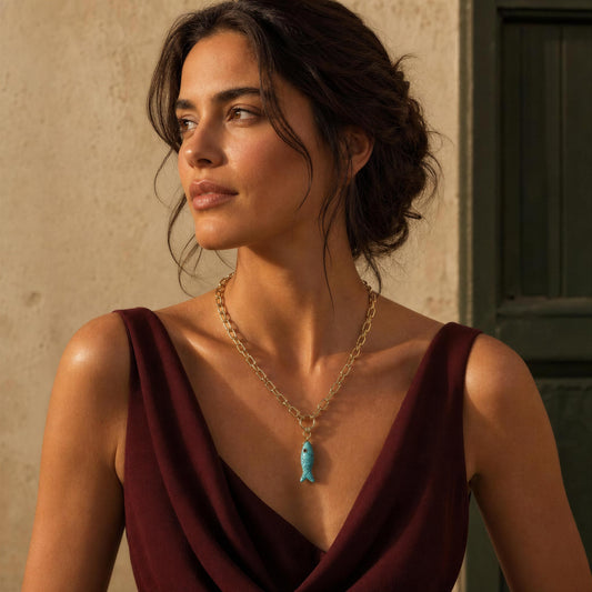 A woman wearing a gold chain necklace with a small turquoise fish pendant, styled with a deep burgundy draped top against a warm beige wall.