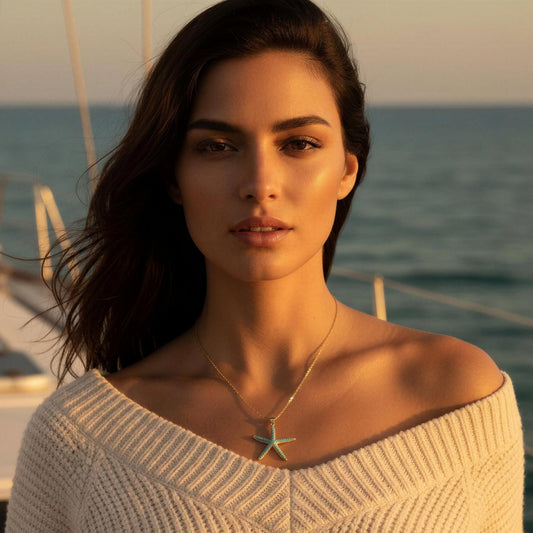 A woman with dark hair stands on a boat with the ocean in the background, wearing a cream off-the-shoulder knit sweater and a delicate gold necklace with a turquoise starfish pendant.