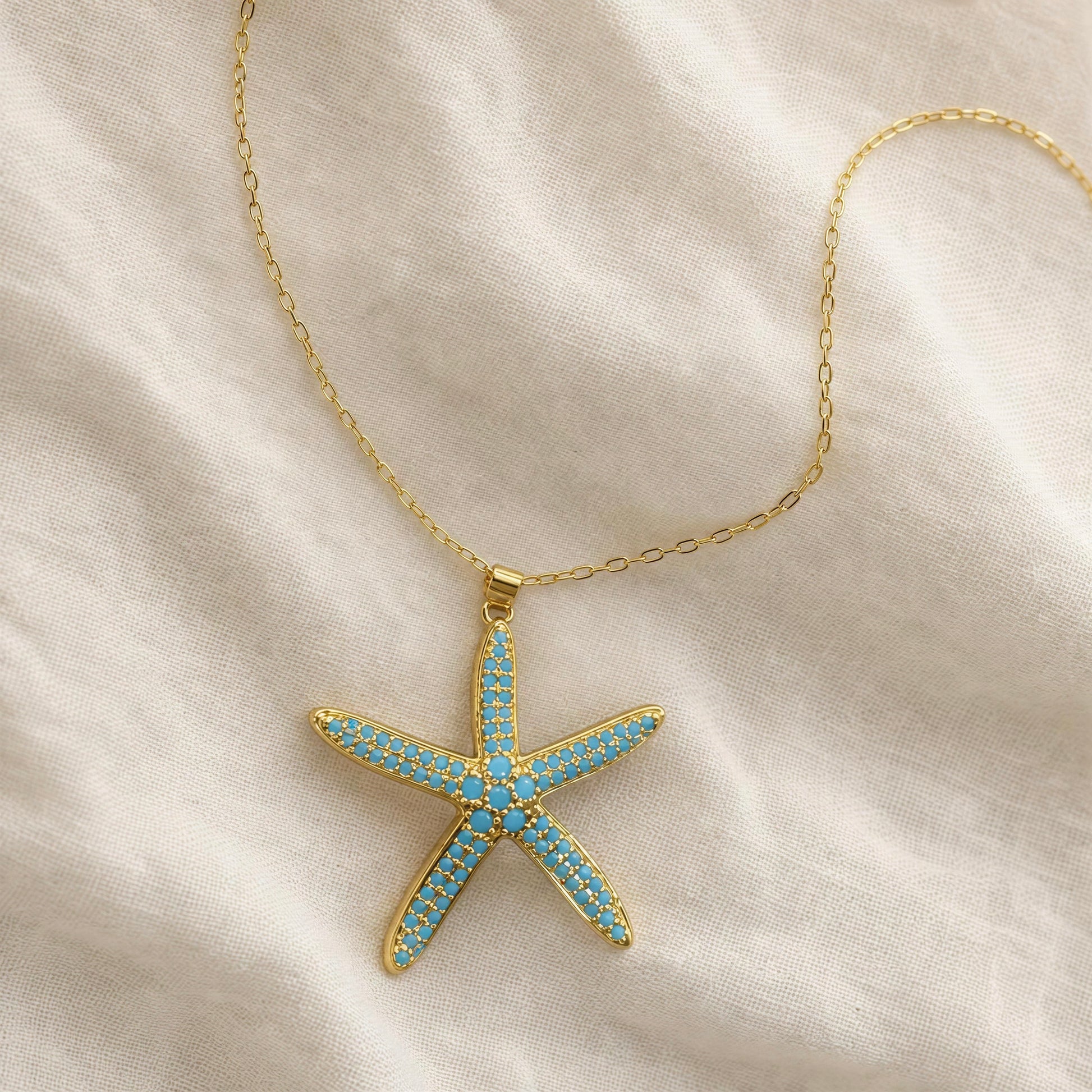 The Asteria Necklace from Chica Chica Jewelry, a gold starfish pendant necklace adorned with light blue rhinestones, displayed on a soft, draped cream-colored fabric background.