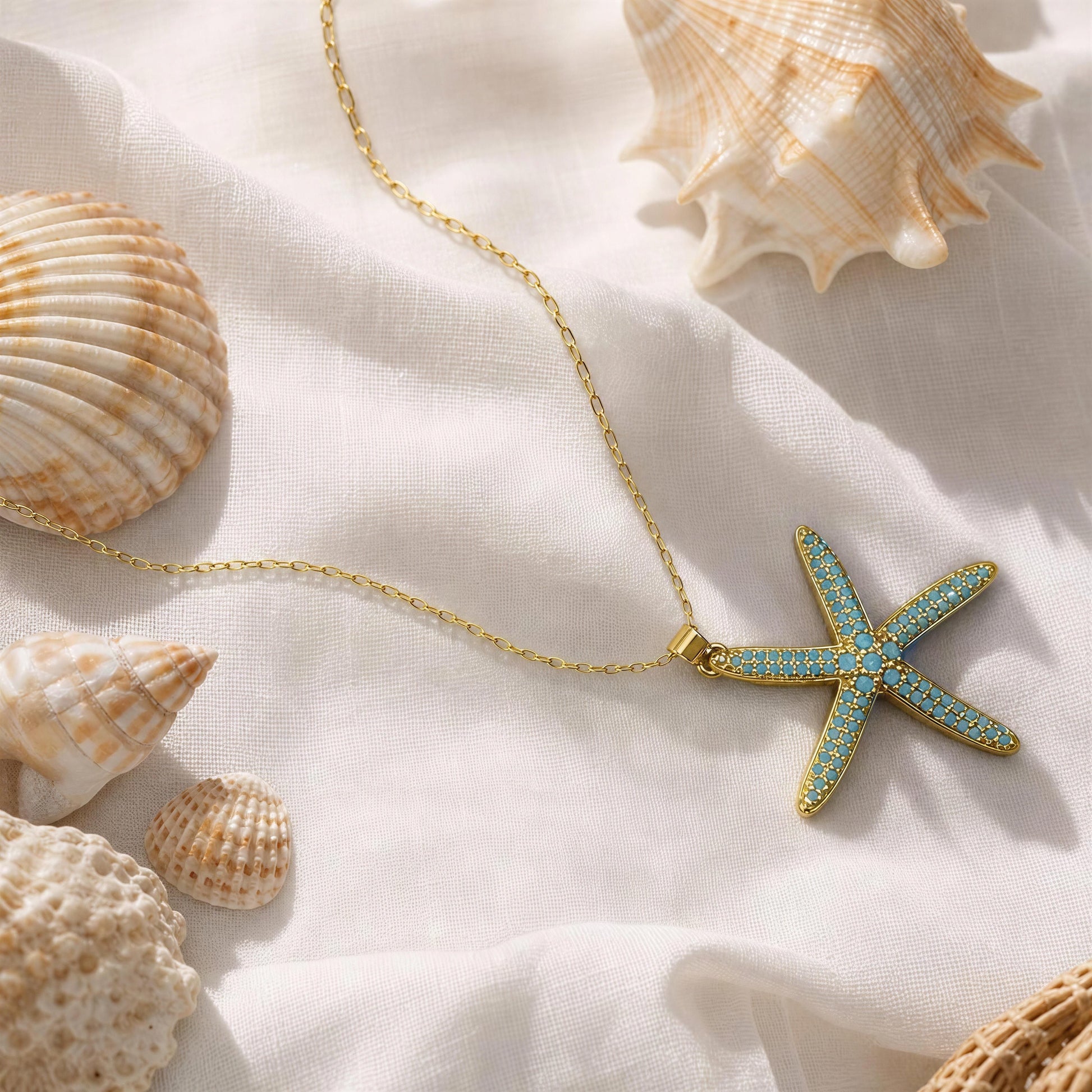 A gold chain necklace with a starfish-shaped pendant featuring turquoise blue and white patterned details, styled on draped white fabric with seashells around it.