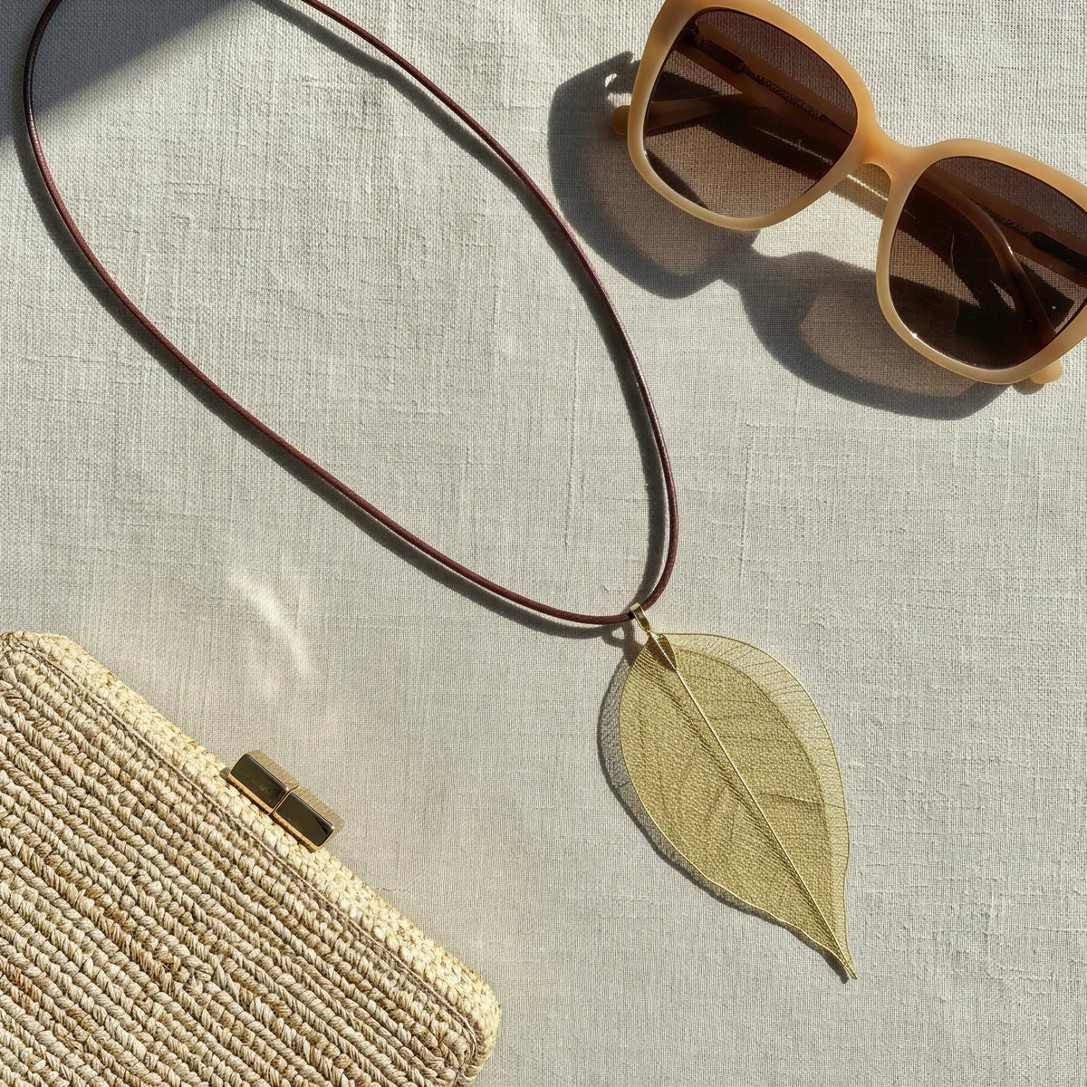 The Aurelia Necklace from  Chica Chica Jewelry placed flat lay on fabric next to sunglasses and a raffia clutch. 