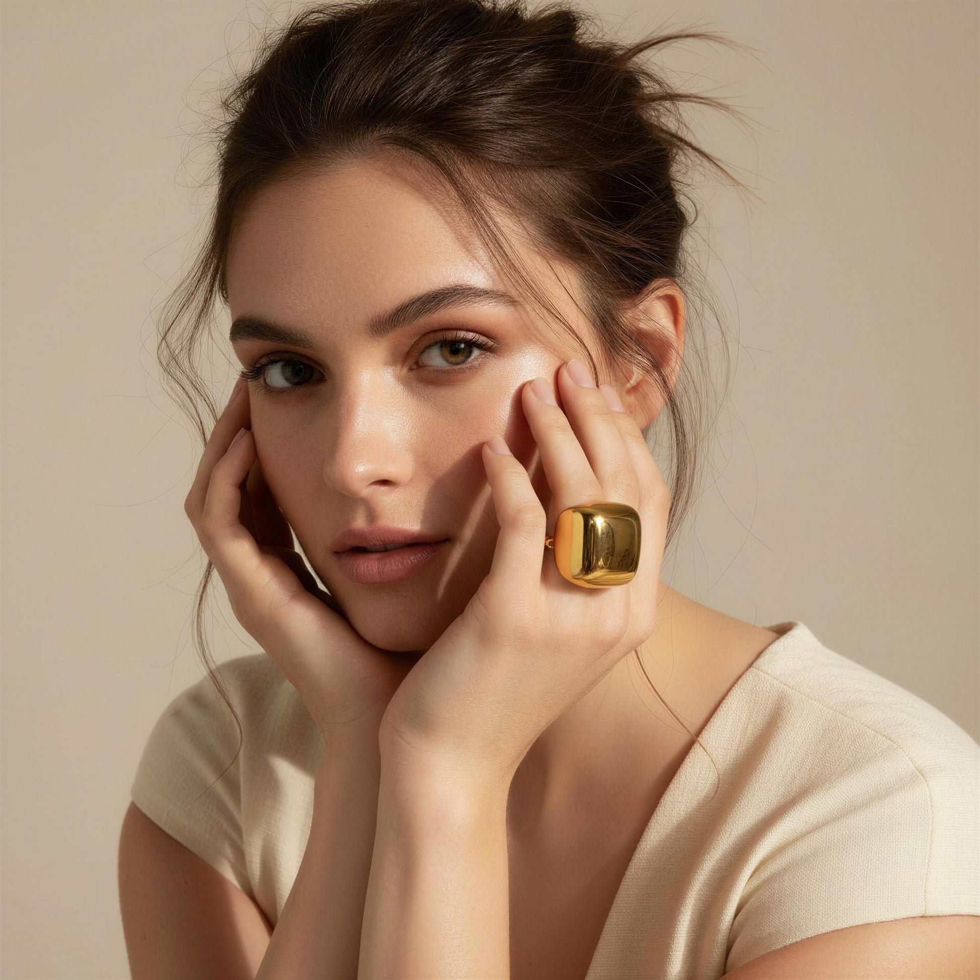 A close-up portrait of a woman with her hands near her face, wearing the Aurelia Ring from Chica Chica Jewelry  on one finger. She is wearing a light beige short-sleeve top against a soft beige background.