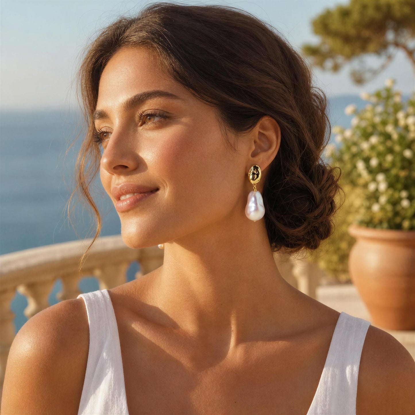 A woman wearing baroque pearl drop earrings, photographed outdoors near the sea with a warm sunny glow.