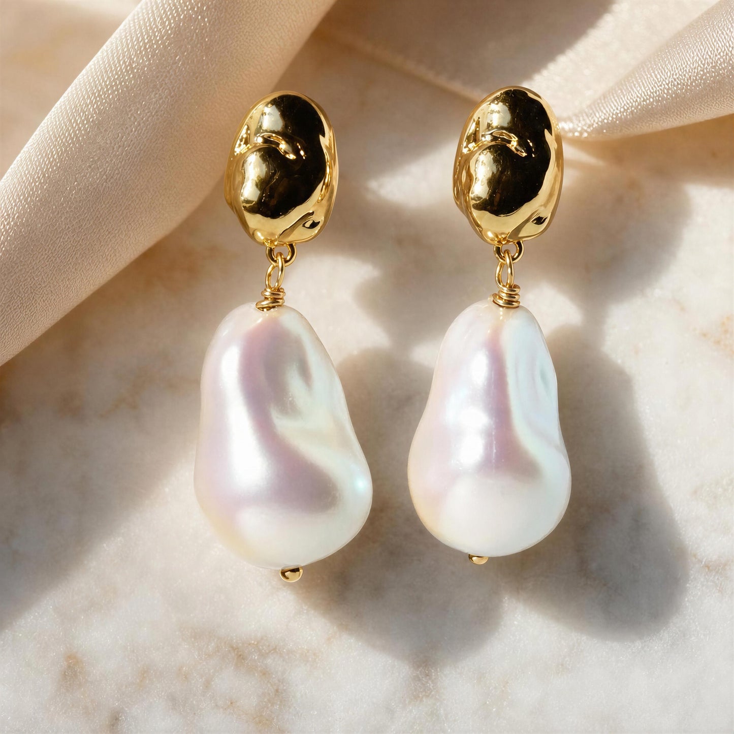 A pair of gold and baroque pearl drop earrings displayed on a soft beige satin ribbon over a light marble surface.