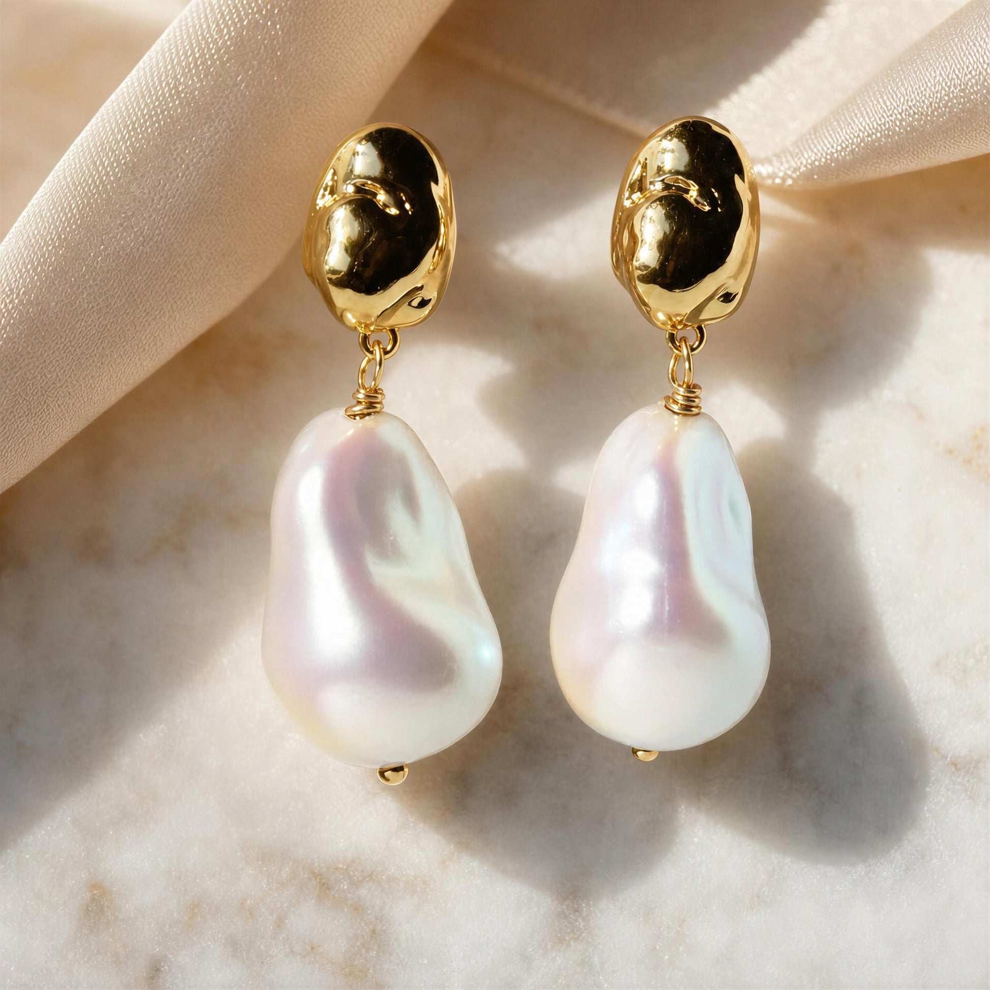 A pair of gold and baroque pearl drop earrings displayed on a soft beige satin ribbon over a light marble surface.