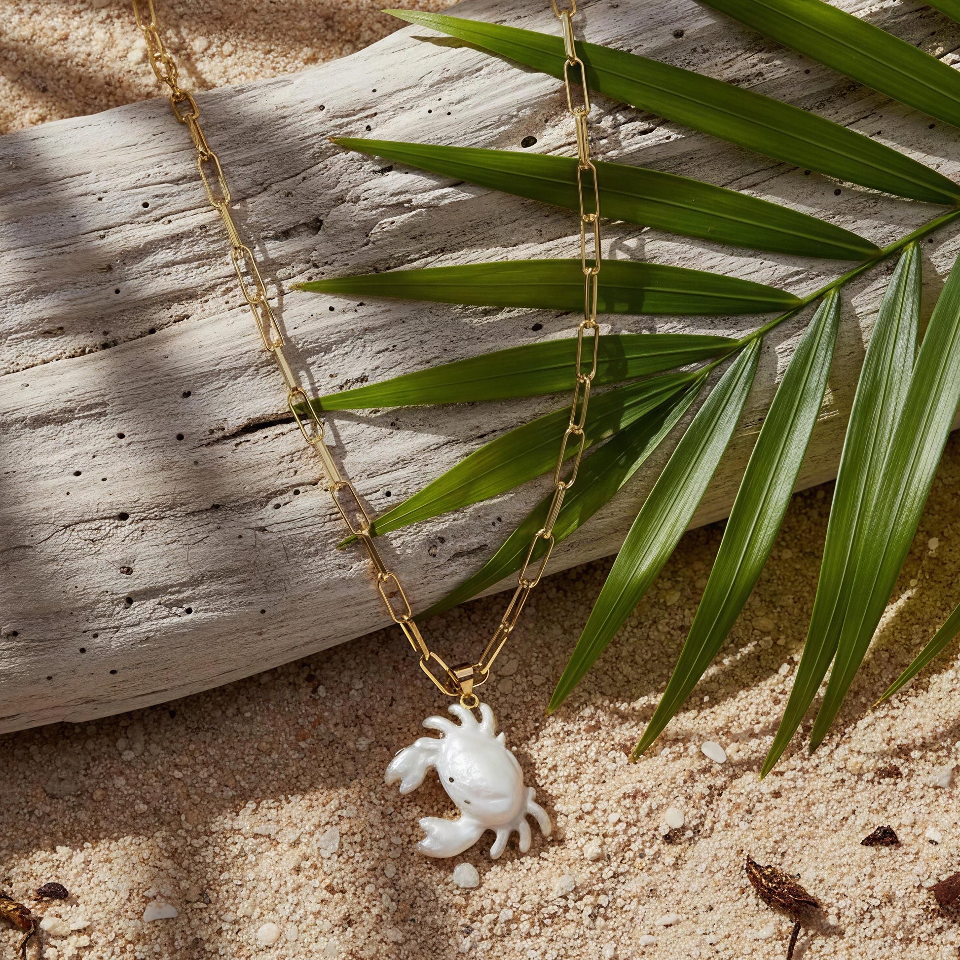 The Brisa Crab Necklace, a gold necklace with a crab pendant on sand and palm leaf. 