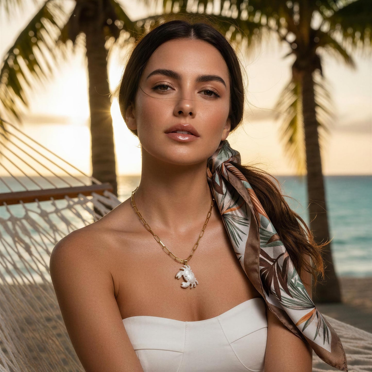 Woman in a white dress with a scarf and necklace, standing in a tropical setting with palm trees and ocean wearing the Brisa Crab Necklace.
