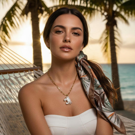 Woman in a white dress with a scarf and necklace, standing in a tropical setting with palm trees and ocean wearing the Brisa Crab Necklace.