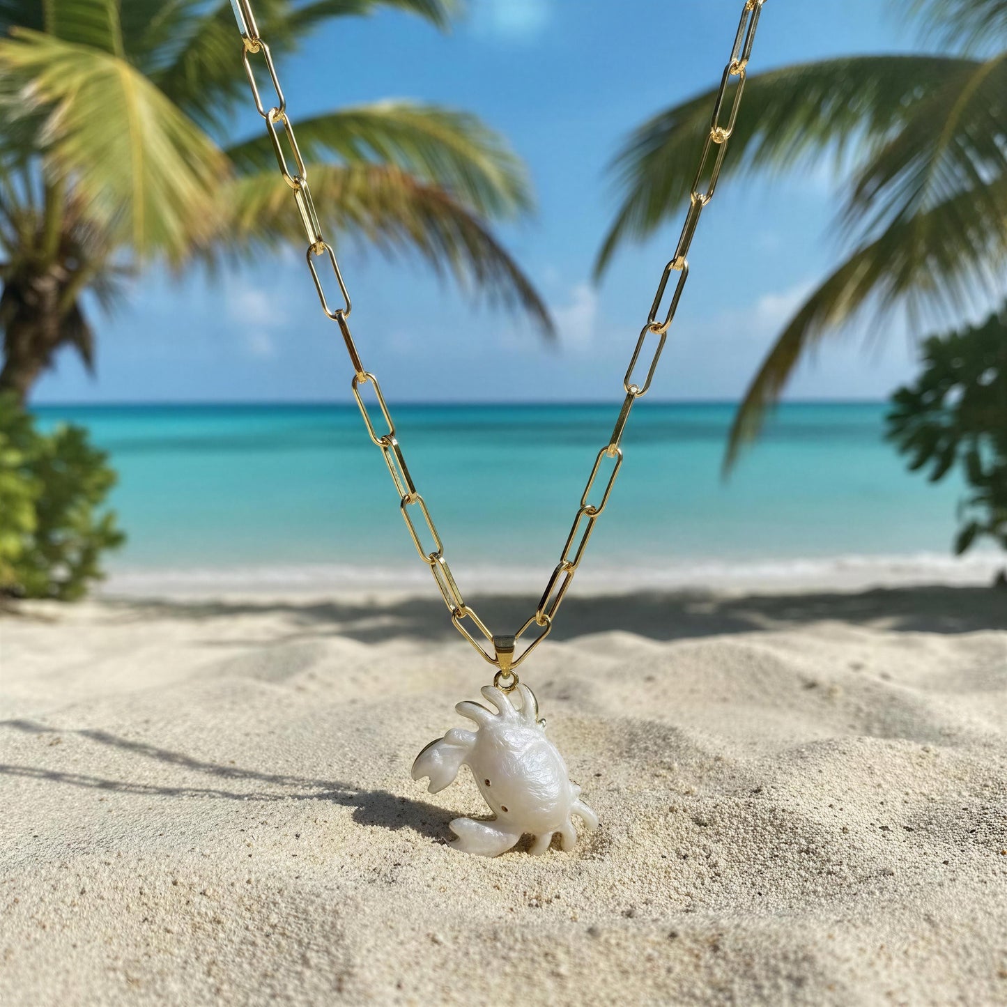 Gold chain necklace with a white crab pendant from Chica Chica Jewelry on a sandy beach with palm trees and blue water in the background.