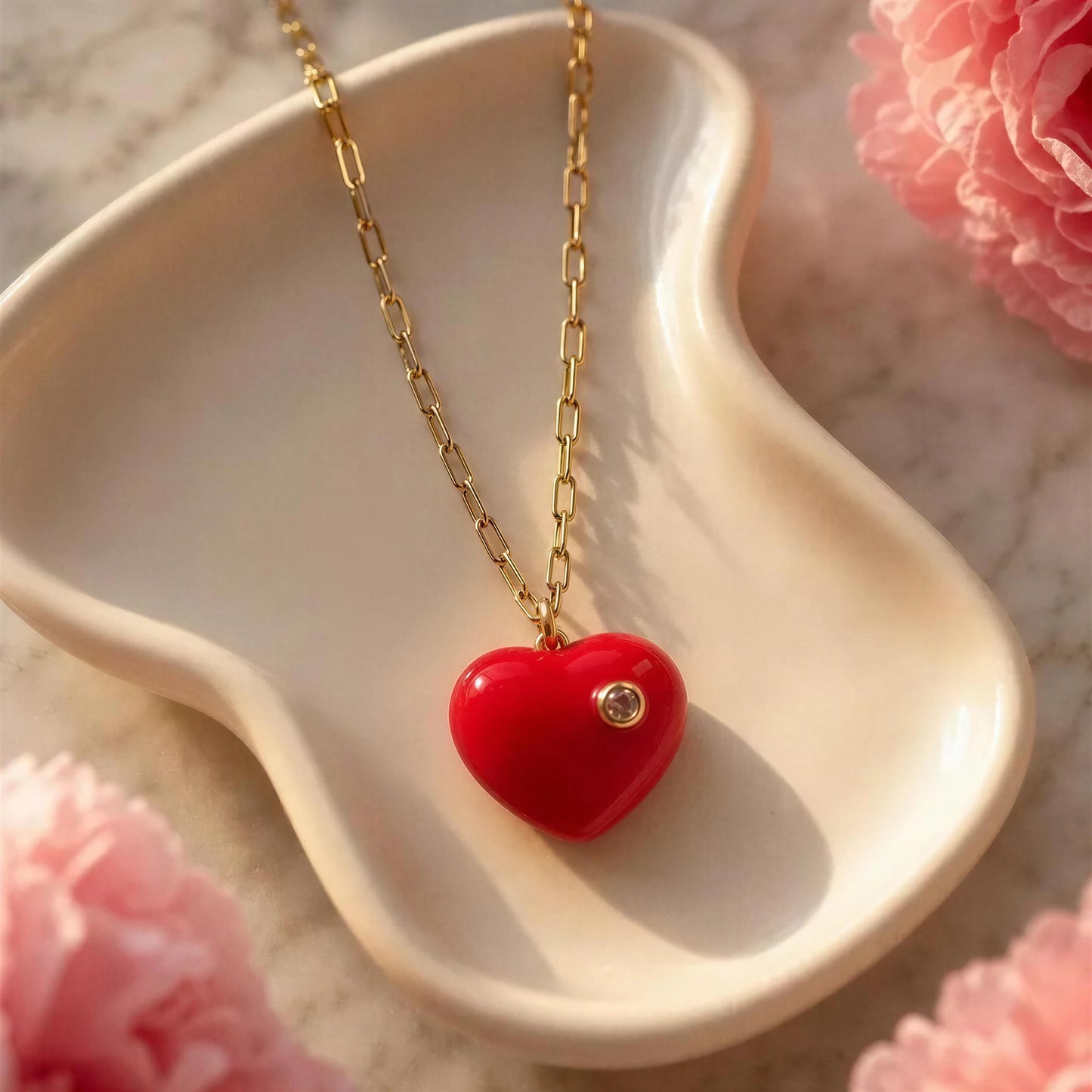 The Amore Fuchsia Heart Necklace on a gold chain, displayed on a heart-shaped white dish with pink flowers in the background.