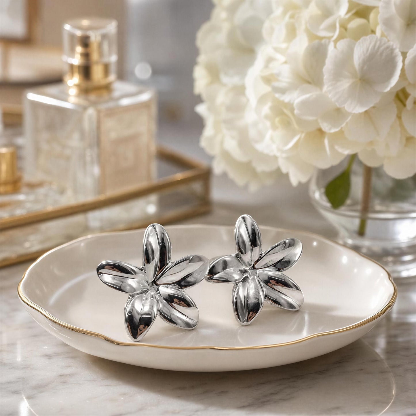 Silver flower-shaped earrings on a white dish with a blurred background of flowers and a perfume bottle.