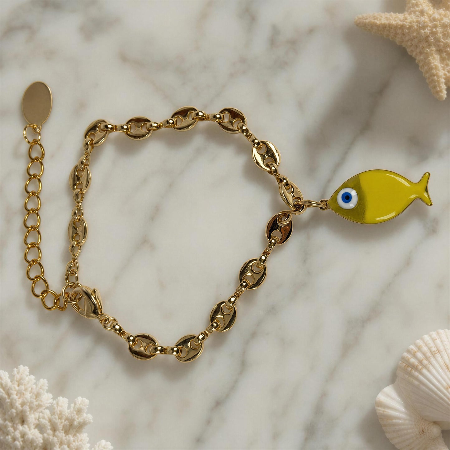 Gold bracelet with a yellow fish charm on a marble surface with starfish and shell decorations.