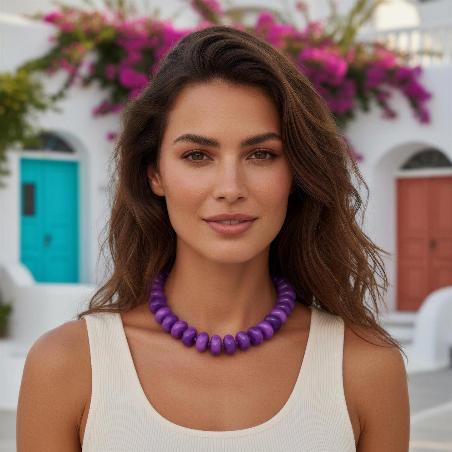 A woman with wavy brown hair wearing the Capri Lilla Choker, a chunky purple beaded necklace and a cream ribbed tank top, set against a Mediterranean-style background with white walls, colorful doors, and pink bougainvillea flowers.