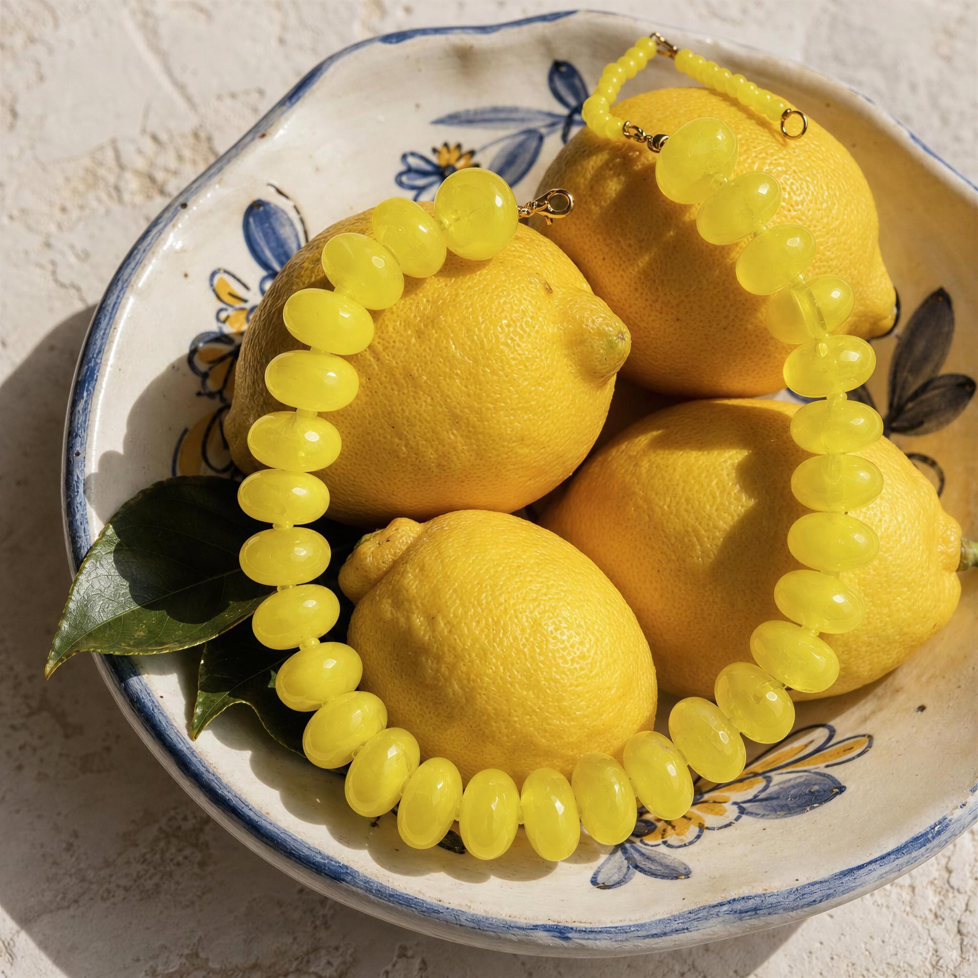 The Capri Limone Choker, from Chica Chica Jewelry draped over several yellow lemons in a white ceramic bowl with blue floral detailing.