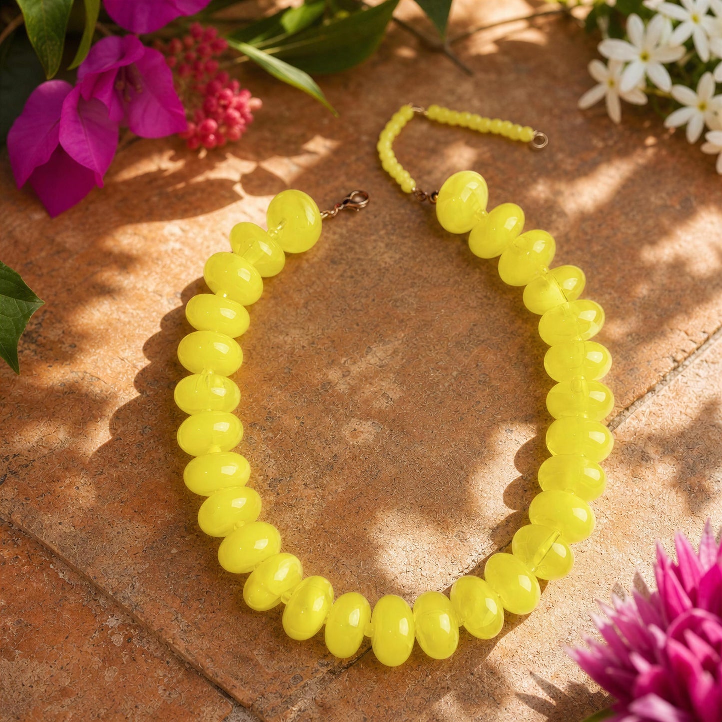 A bright yellow beaded necklace with oval beads and a matching yellow cord clasp, arranged on a terracotta tile surface with flowers in the corners.
