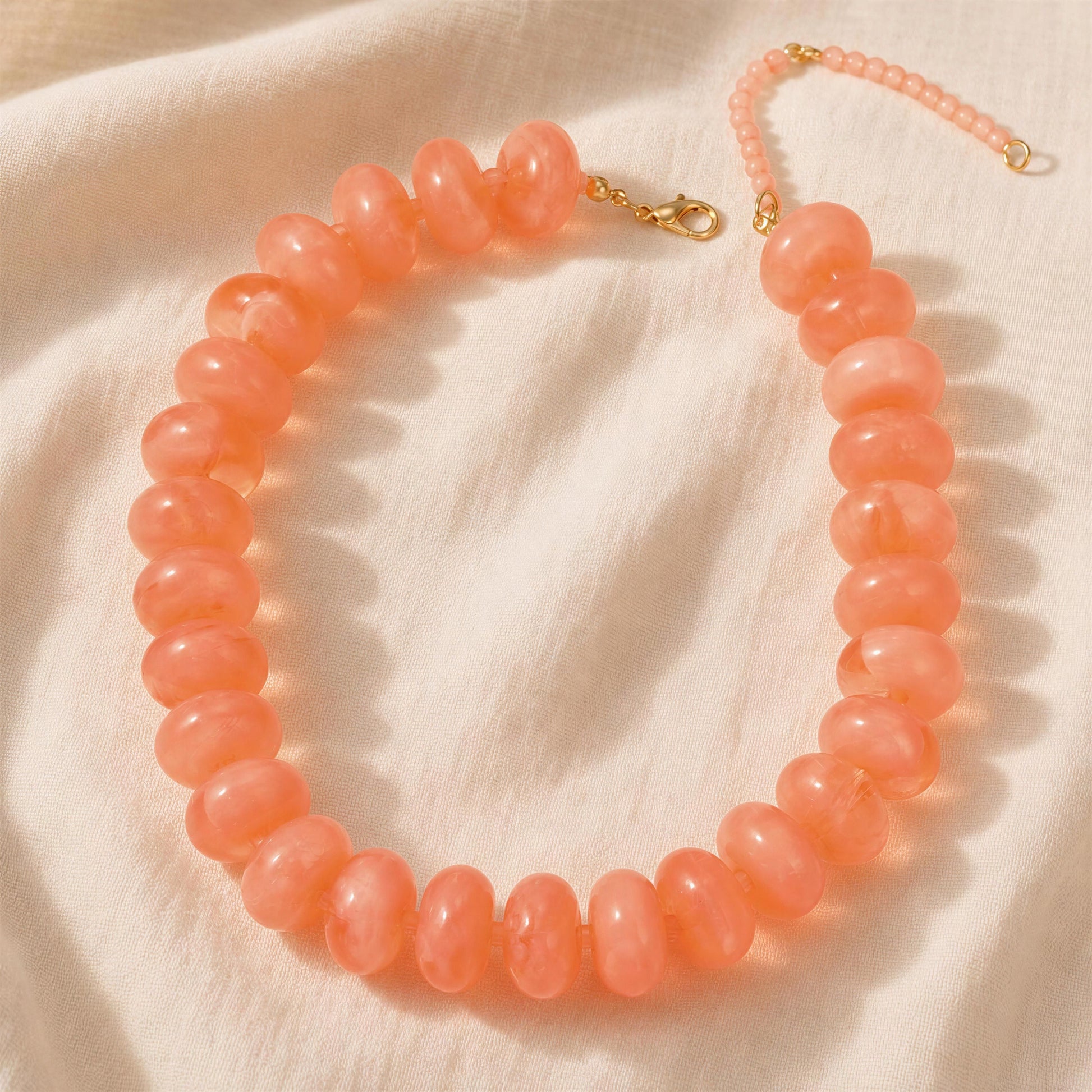 The Capri Pesca Choker, a peach-orange beaded necklace with smooth, rounded oval beads and a gold-tone clasp, displayed on a textured off-white fabric background.