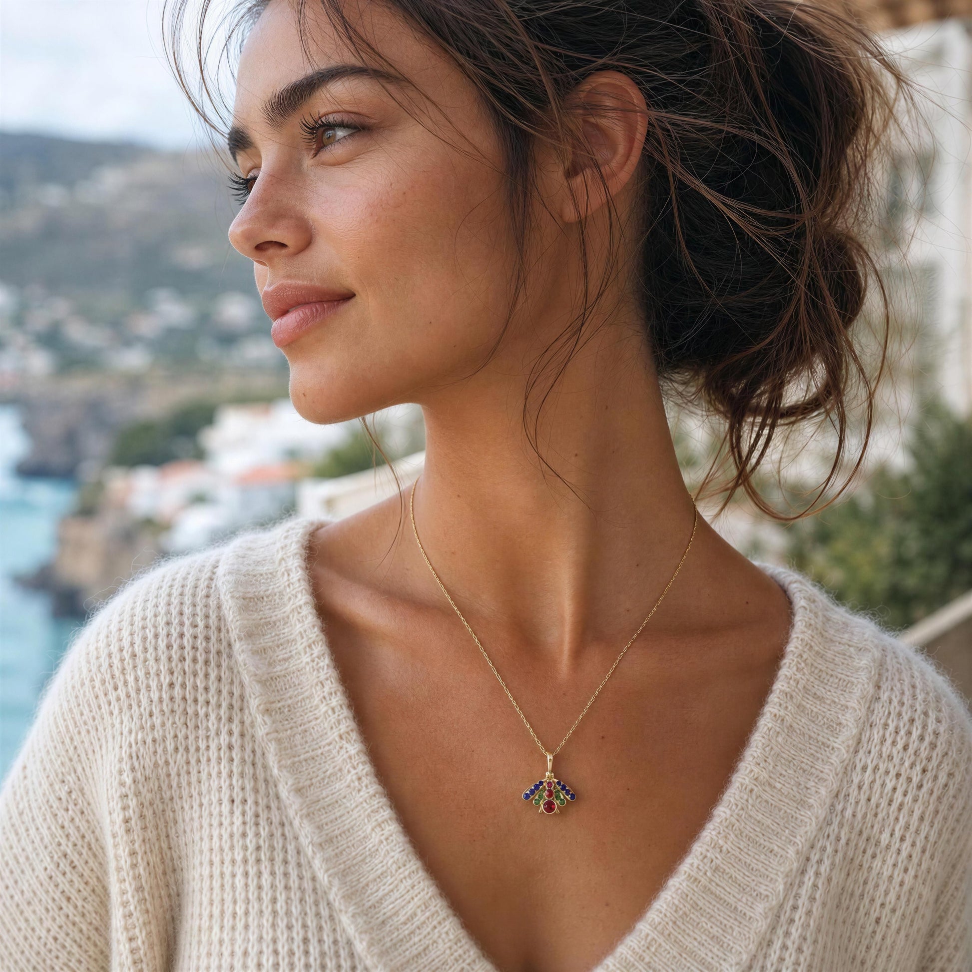 A woman in a cream knit sweater wearing a delicate gold chain necklace with a small, colorful gemstone pendant, set against a blurred coastal background.