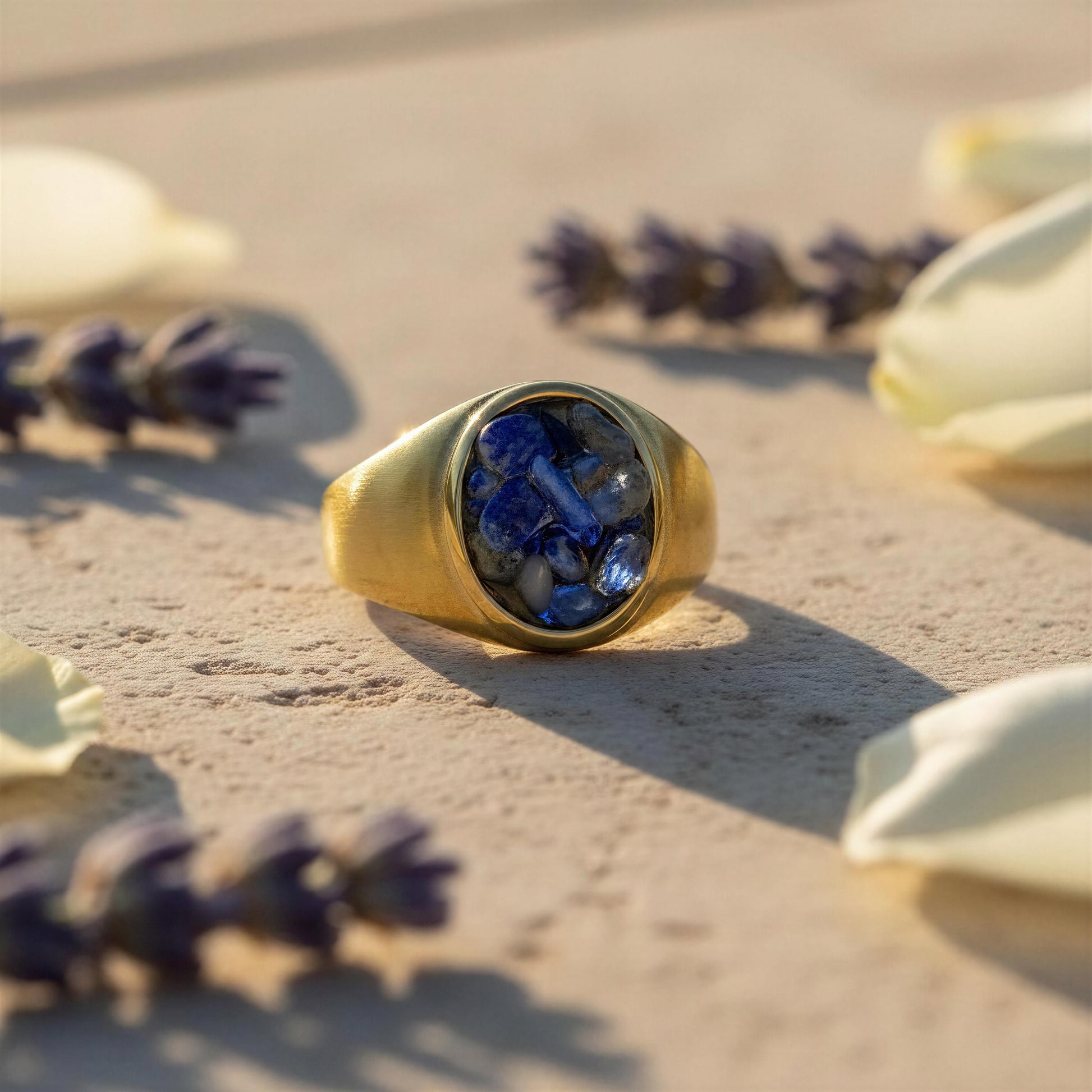 A gold signet-style ring with a deep blue textured stone inlay, resting on a sandy surface surrounded by blurred lavender buds and cream petals.