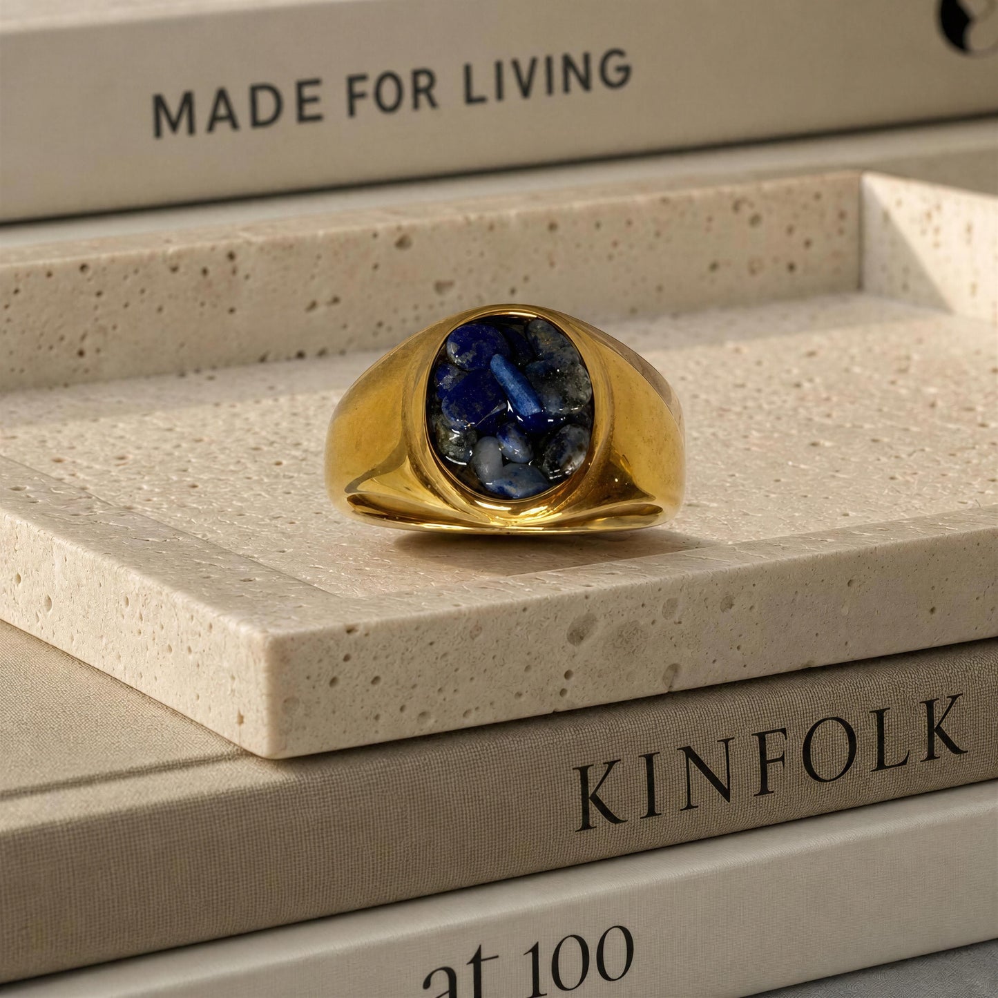 A gold signet ring with a deep blue oval stone, displayed on a textured beige stone slab resting on books.