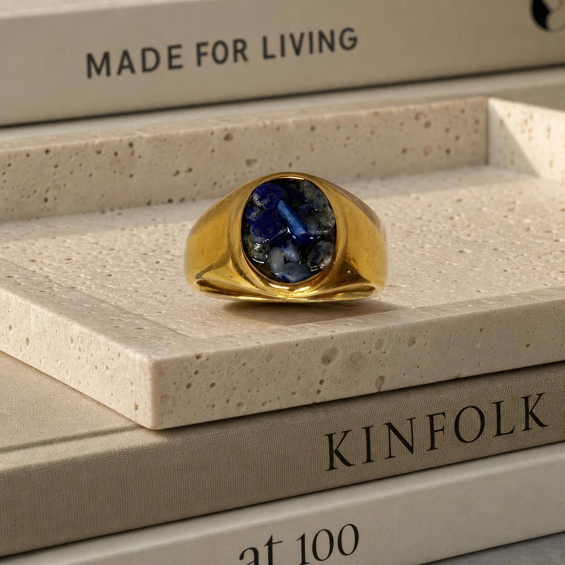 A gold signet ring with a deep blue oval stone, displayed on a textured beige stone slab resting on books.