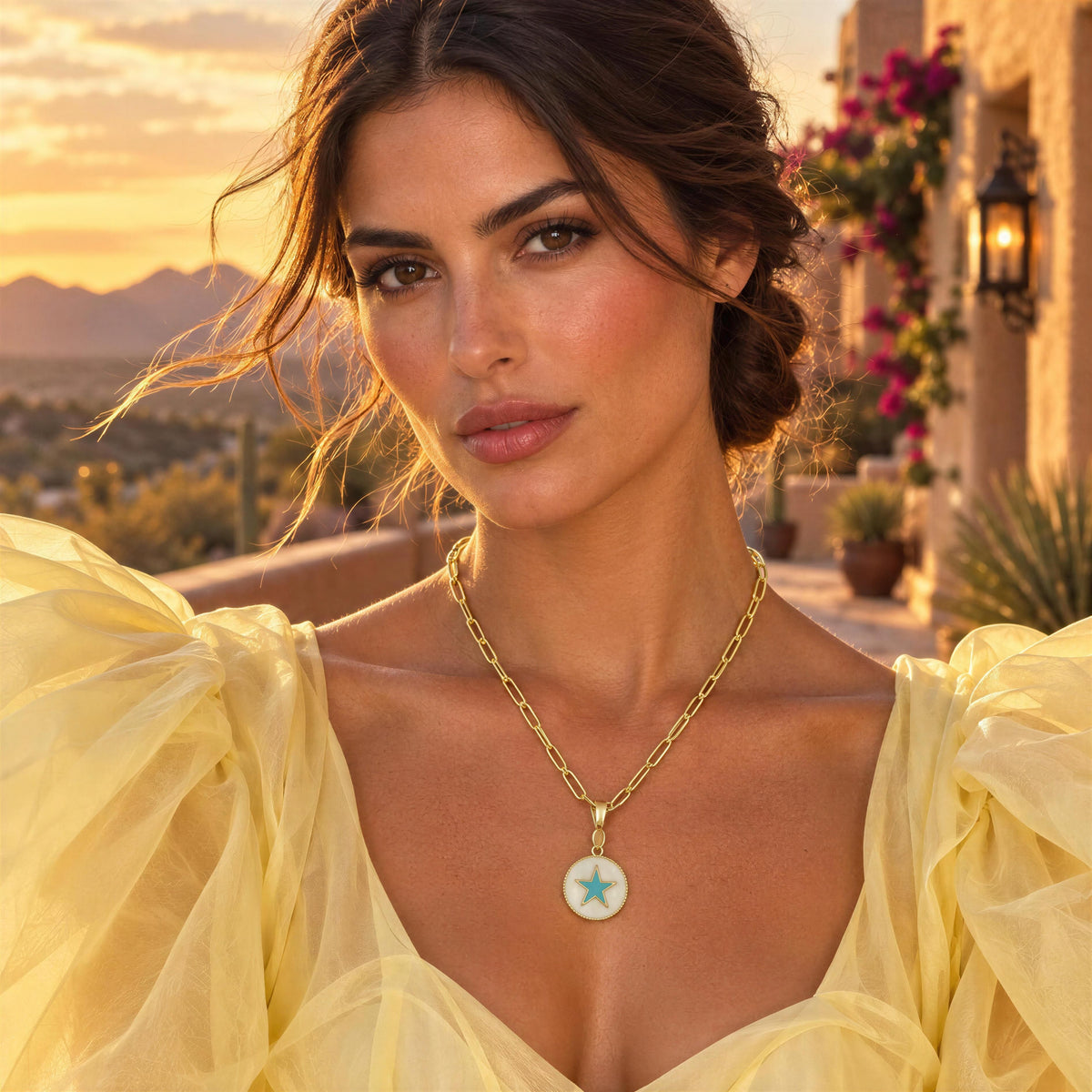 A woman wearing the Celestia Turquoise Star Necklace from Chica Chica Jewelry, a gold chain necklace with a round pendant featuring a turquoise star on a white background, photographed outdoors at sunset with mountains and a balcony in the background.