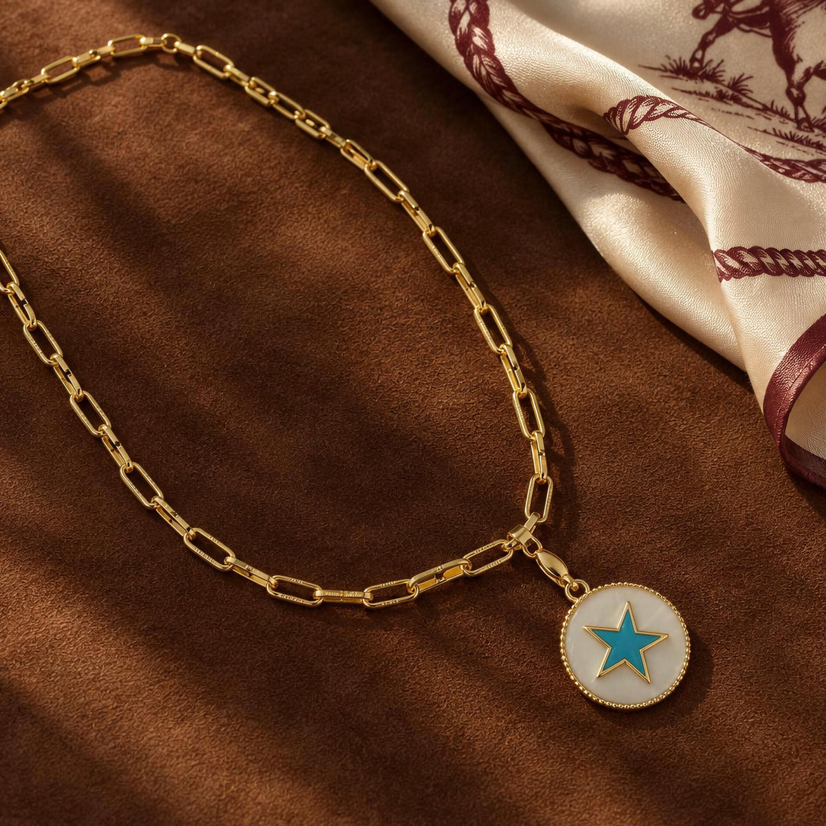 A gold paperclip chain necklace with a round white pendant featuring a blue star, styled on a brown textured surface next to a cream silk scarf.