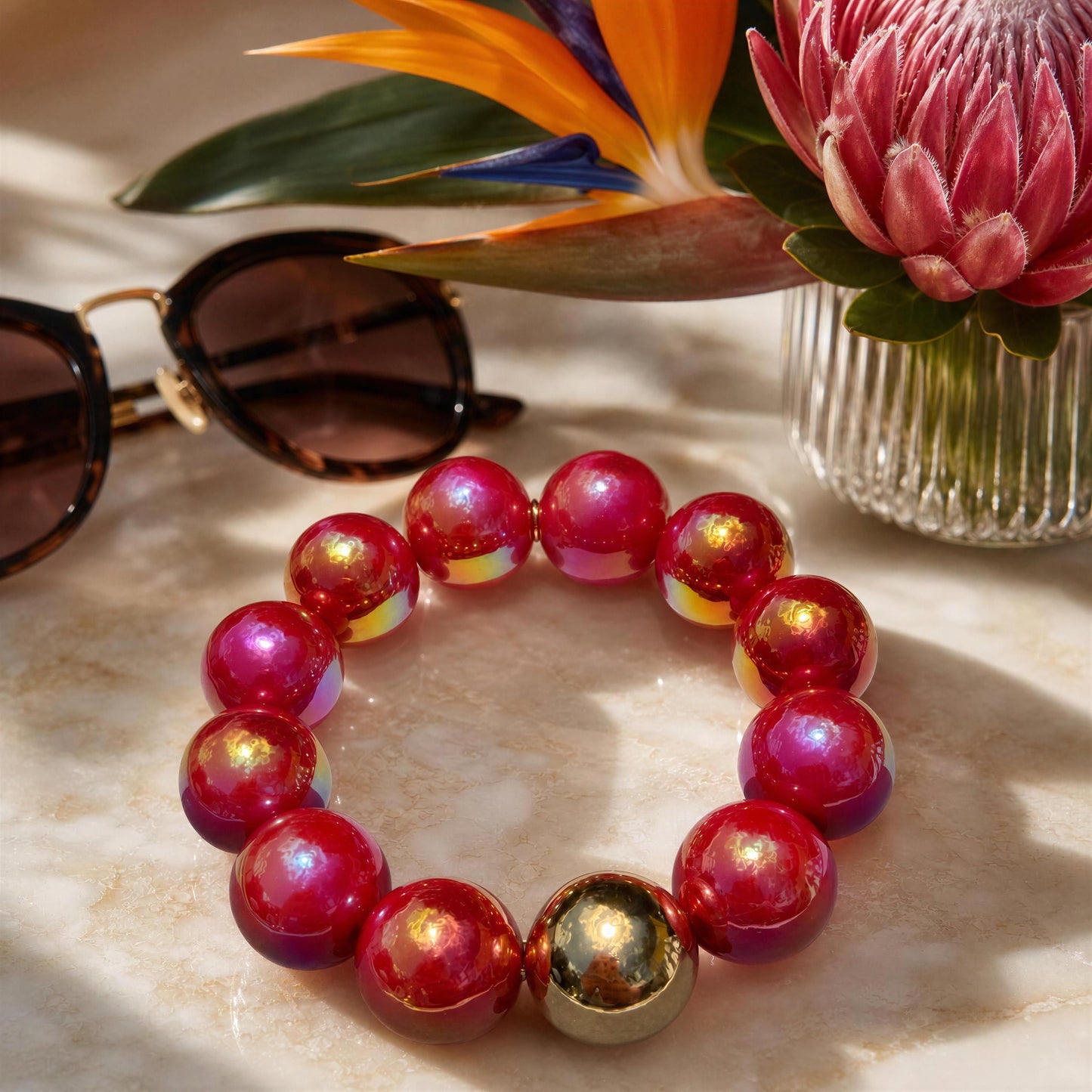 Red beaded bracelet with gold accents on a marble surface with sunglasses and flowers.
