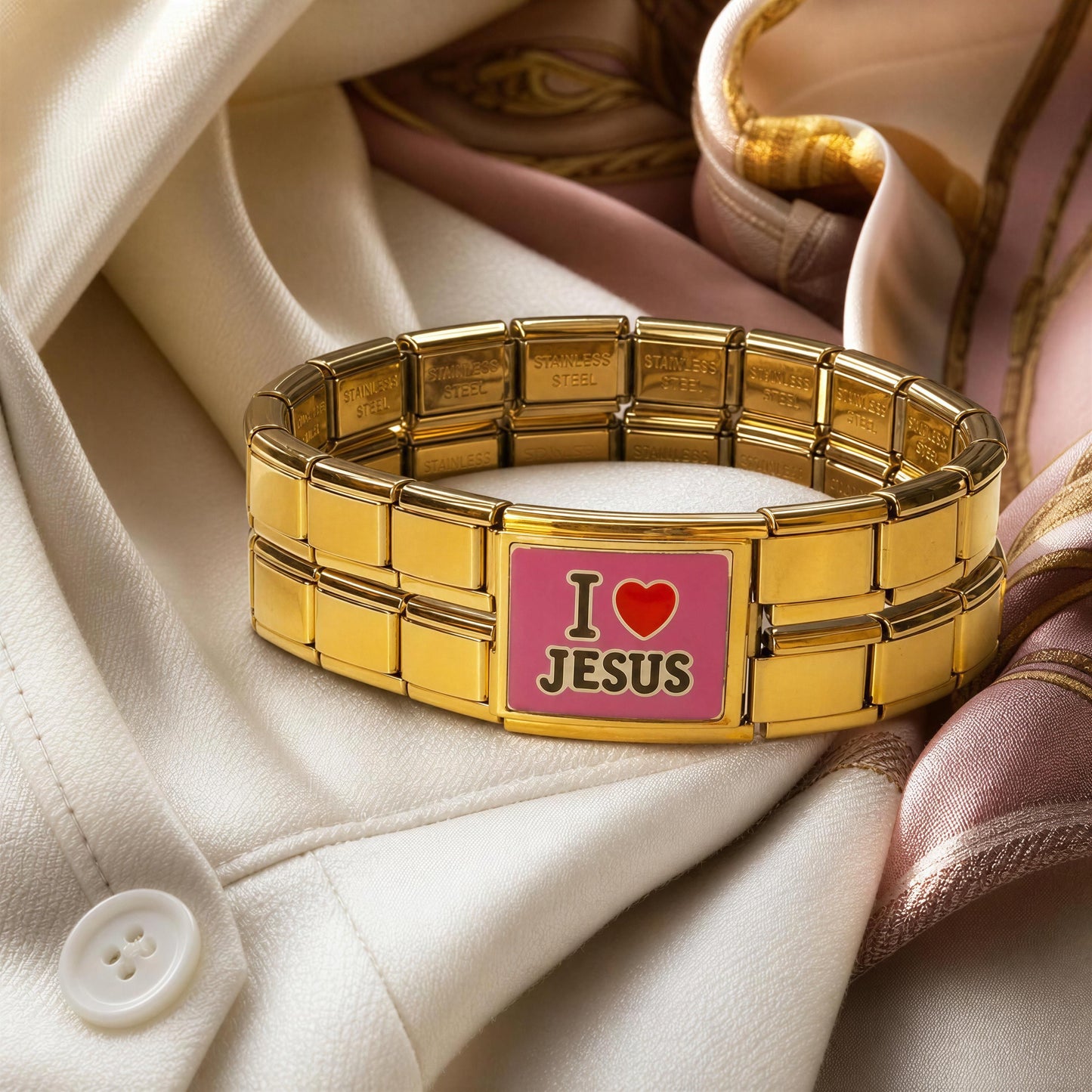 A gold-tone Italian charm bracelet with a rectangular pink clasp that reads 'I love JESUS' in black and red lettering, styled on draped cream and pink fabric.
