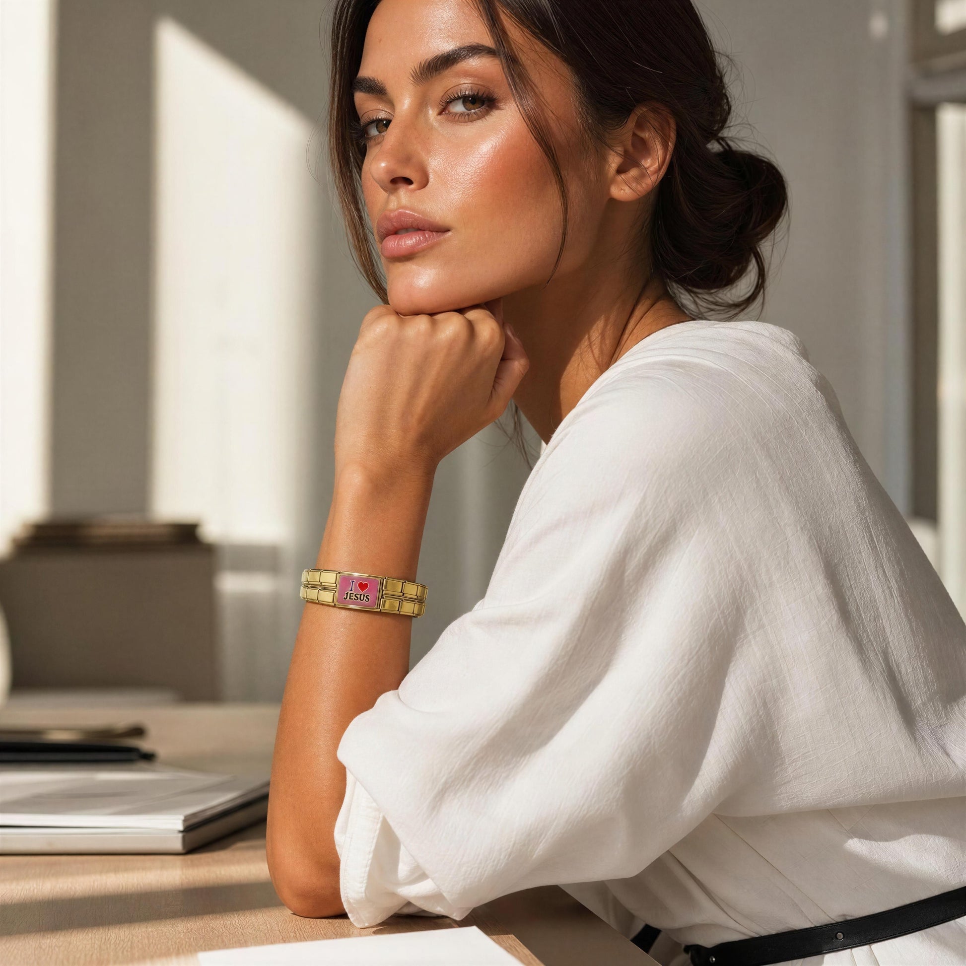 A woman in a white blouse rests her chin on her hand, wearing the Amada Bracelet, a  gold-toned bangle bracelet. The setting is a bright, modern indoor with  soft natural light.