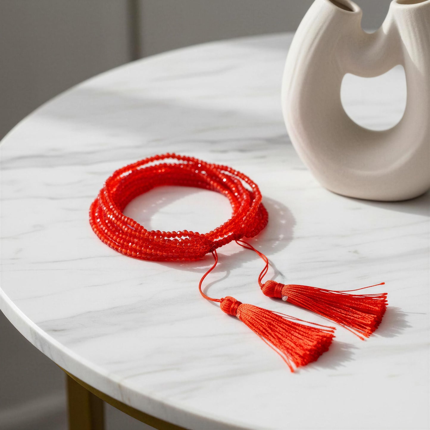 Red beaded bracelet with tassels on a marble surface next to a white vase.
