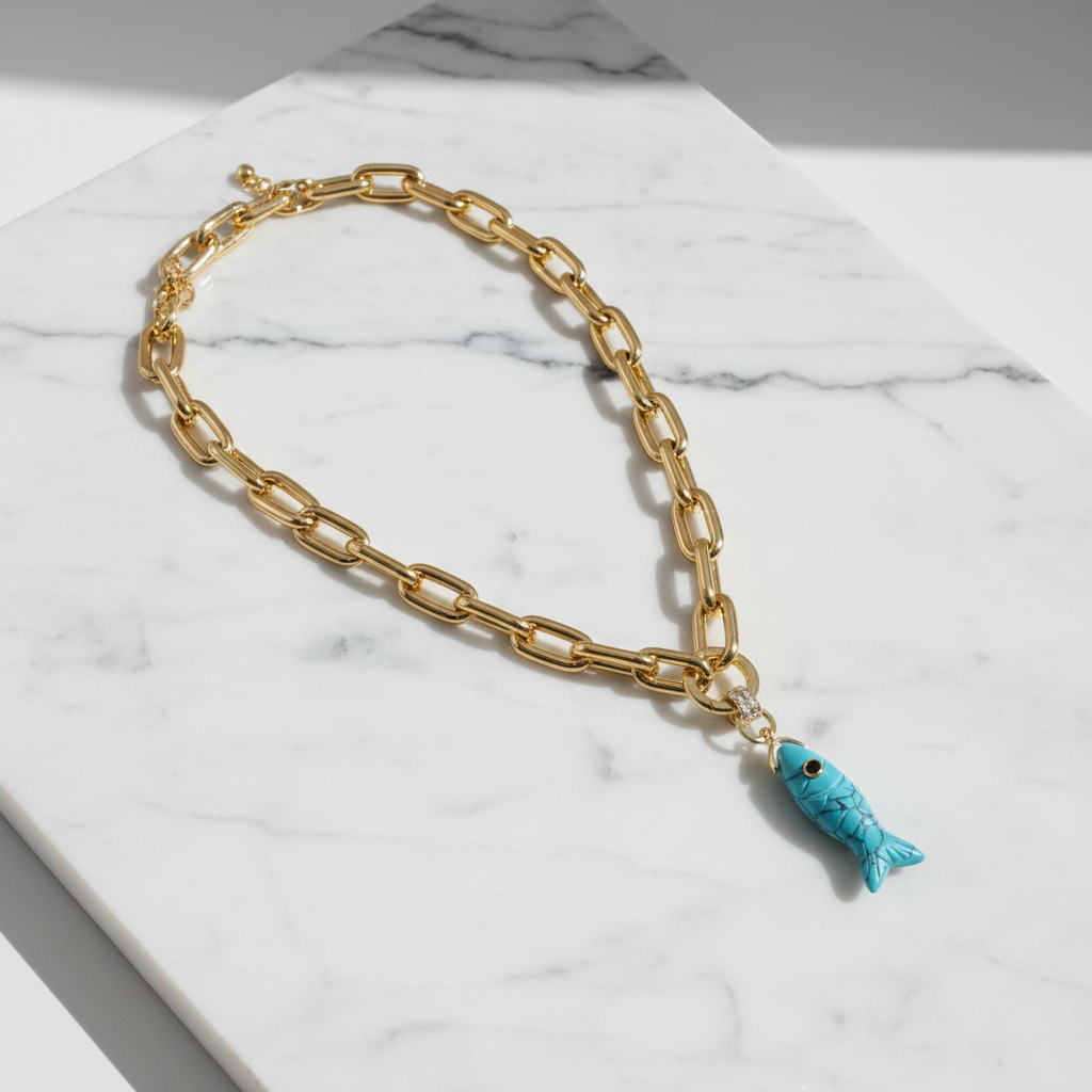 Gold chain bracelet with a turquoise fish charm on a marble surface