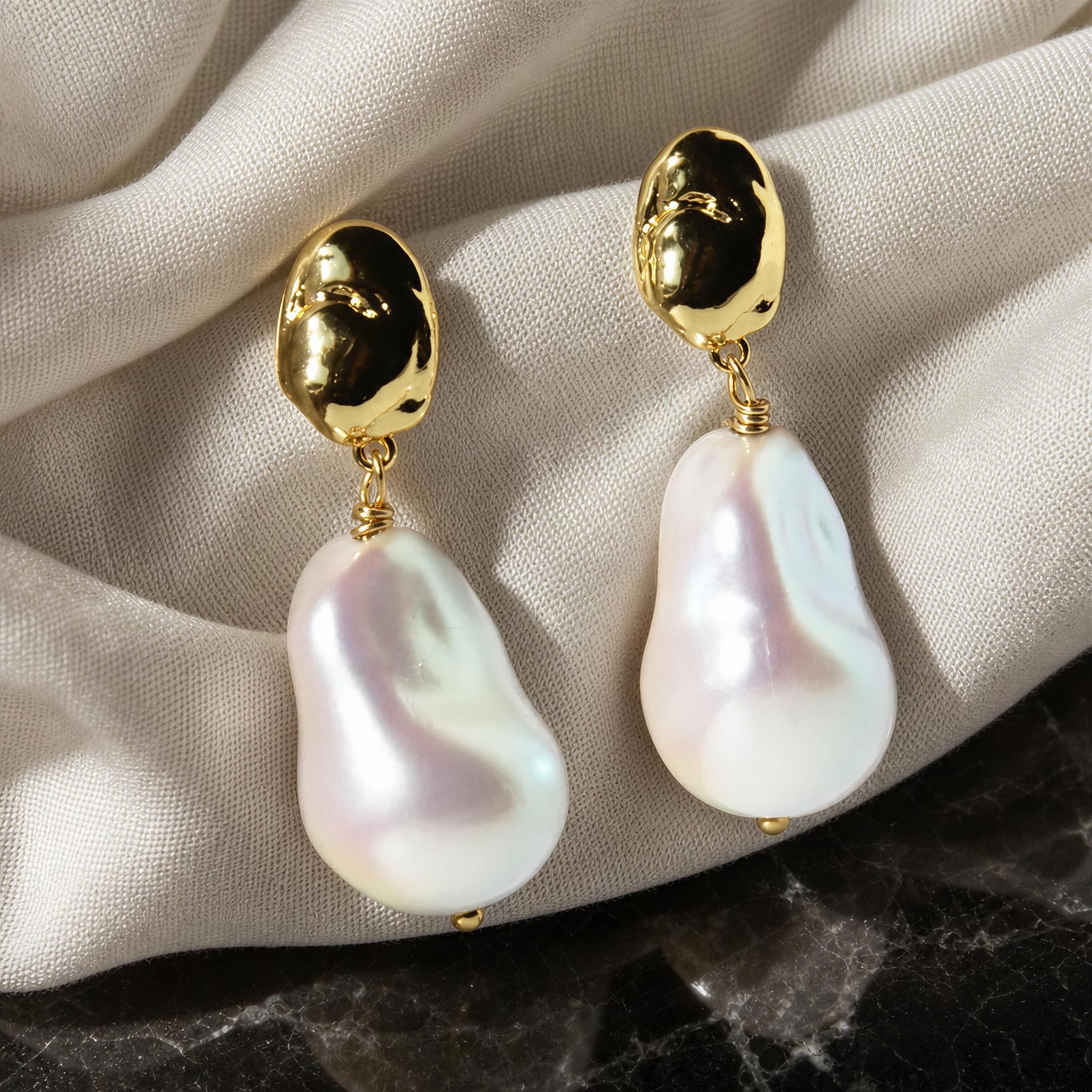 A pair of gold drop earrings with irregular white baroque pearls, displayed on draped beige fabric over a dark marble surface.
