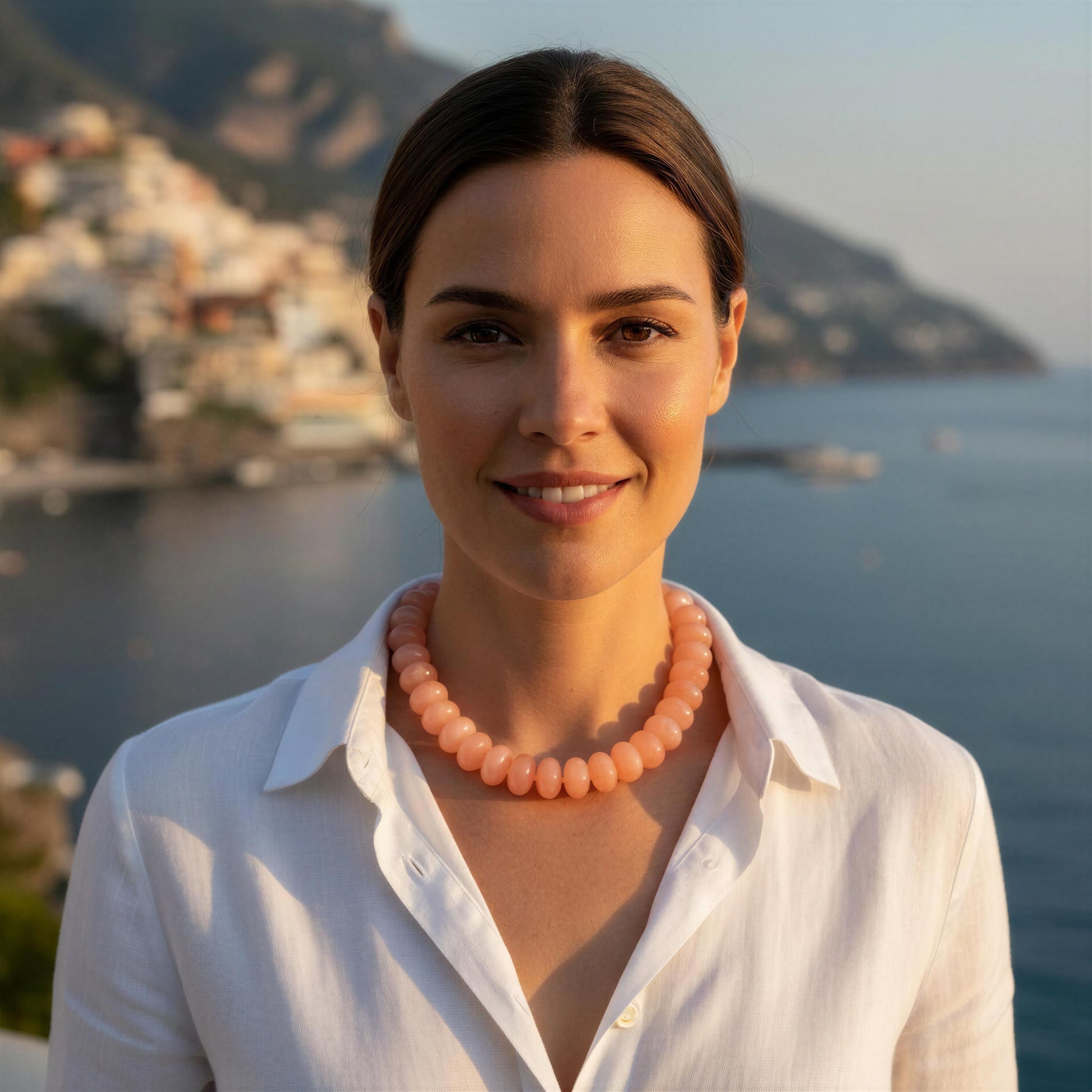 A woman with dark hair pulled back, wearing a white collared shirt and a peach-colored beaded necklace, standing outdoors with a coastal town and sea in the background.