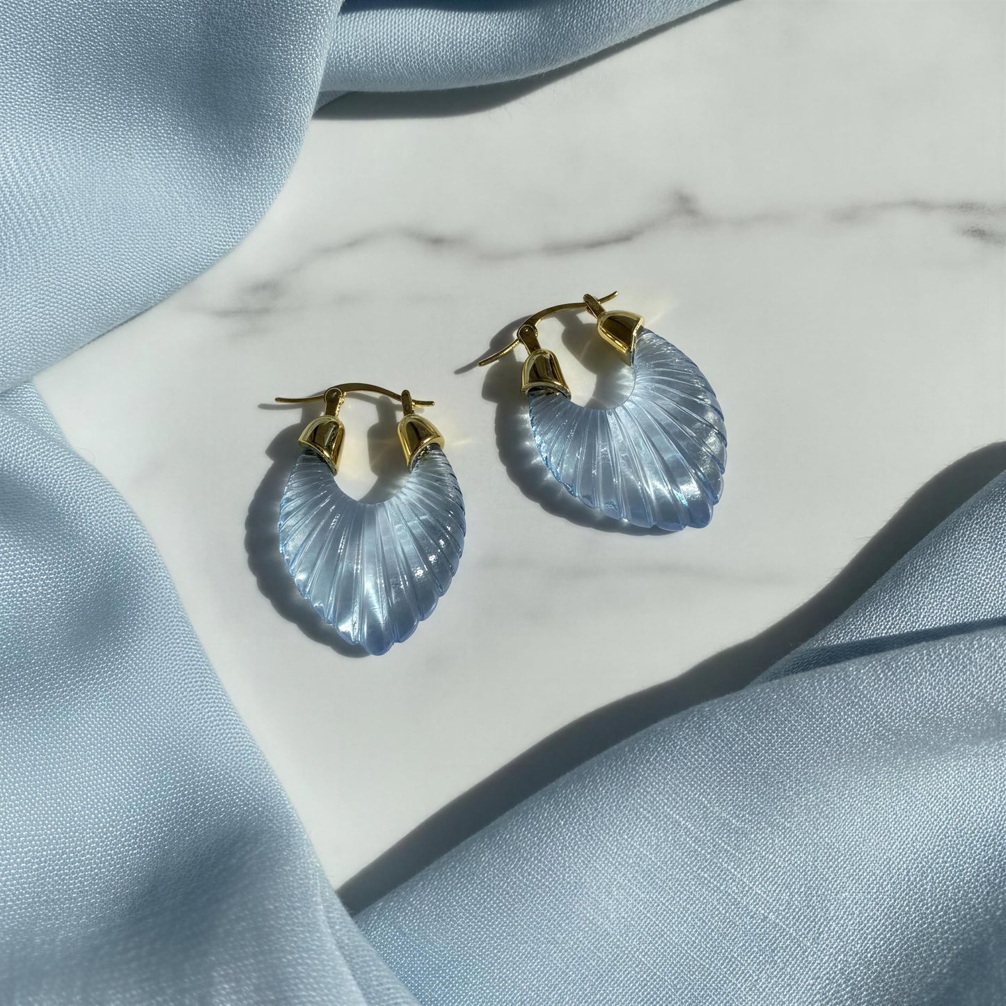 A pair of gold hoop earrings with light blue ribbed crescent-shaped drops, styled on a white marble surface with light blue fabric draping.