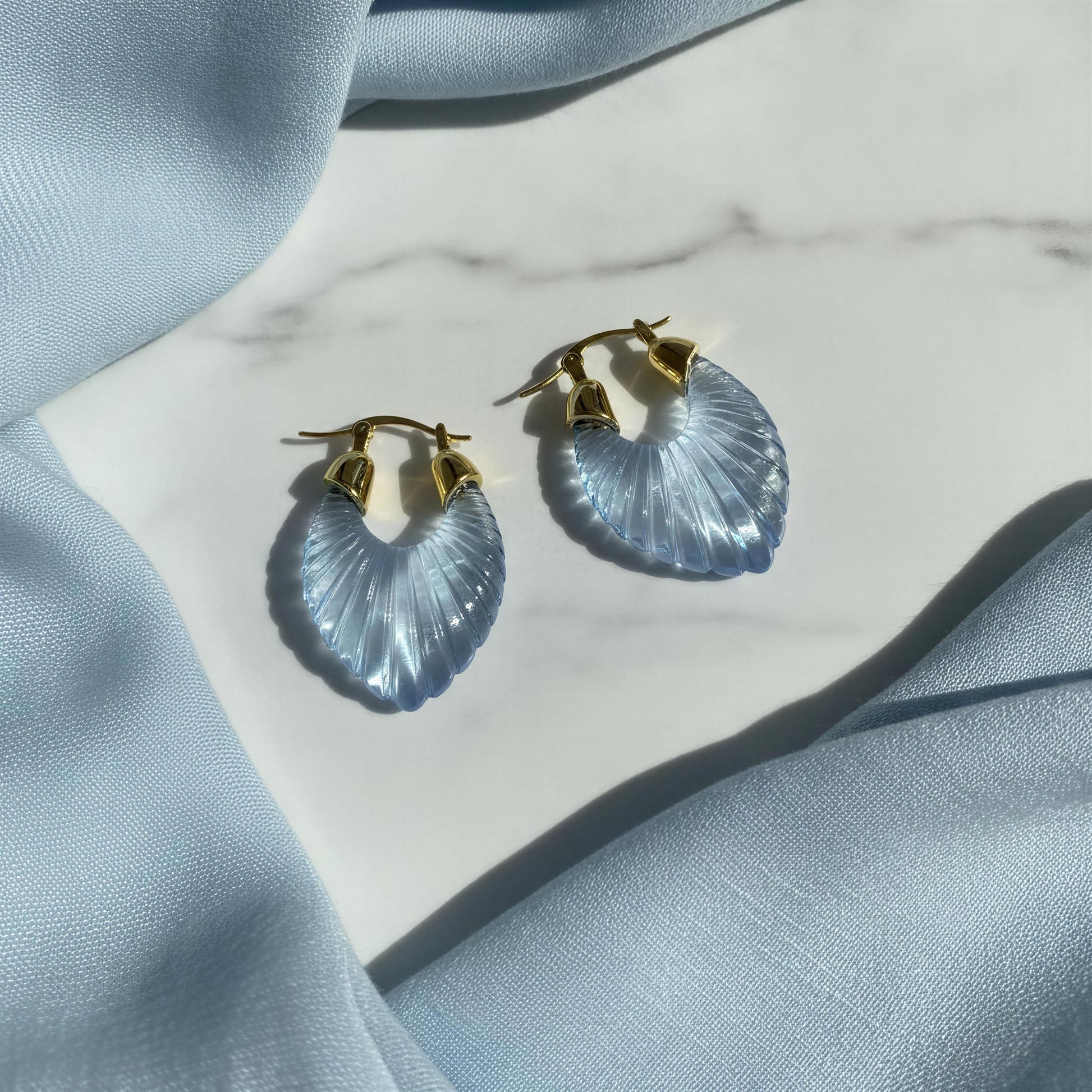 A pair of gold hoop earrings with light blue ribbed crescent-shaped drops, styled on a white marble surface with light blue fabric draping.