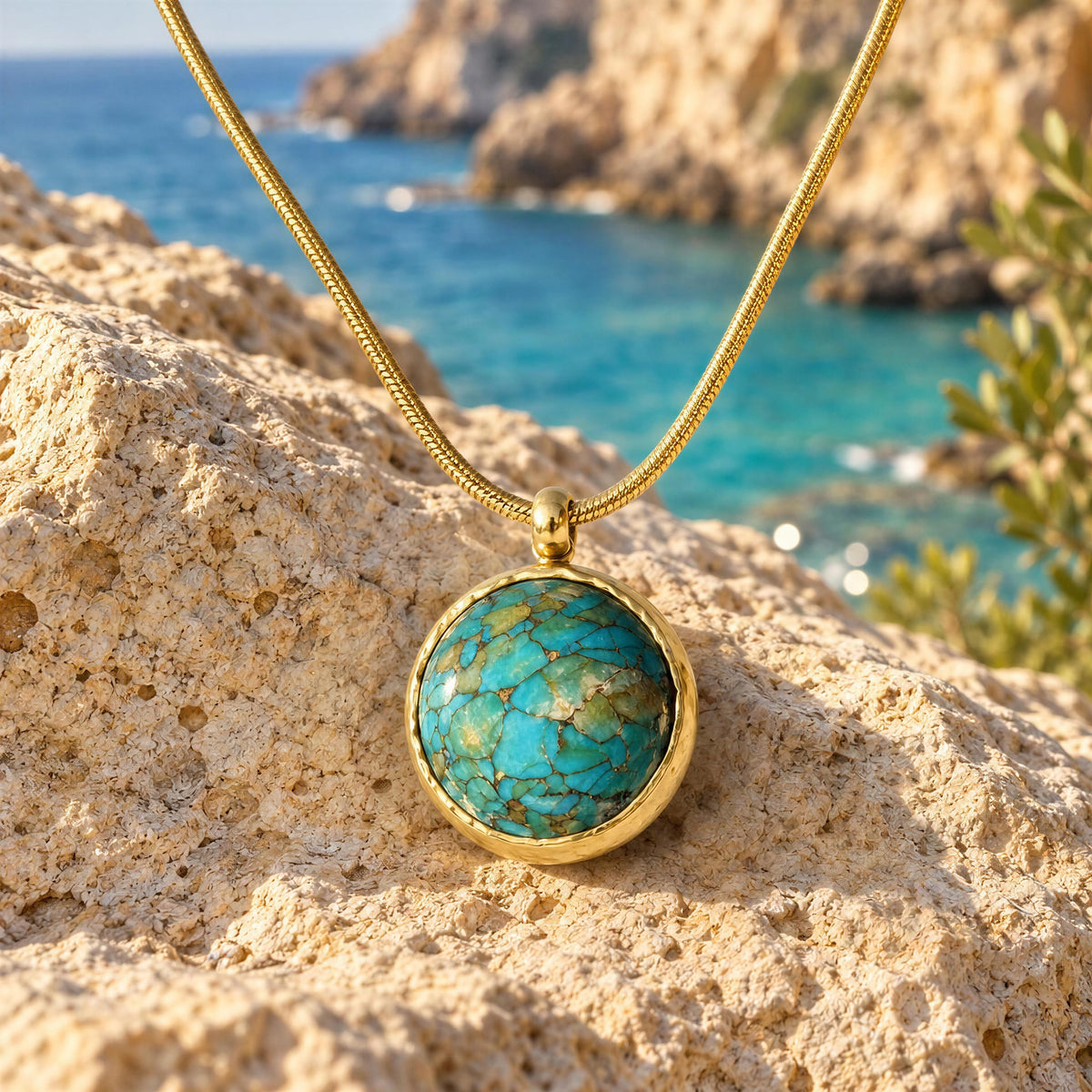 Derya gold-tone necklace with round turquoise blue pendant displayed on natural coastal stone overlooking the sea.