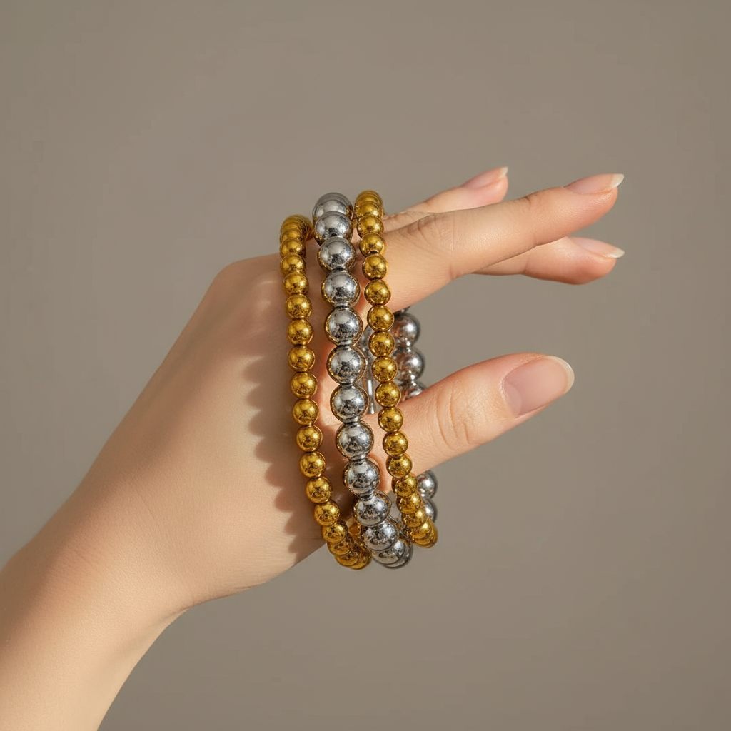 Hand wearing three beaded bracelets in gold, silver, and bronze on a neutral background