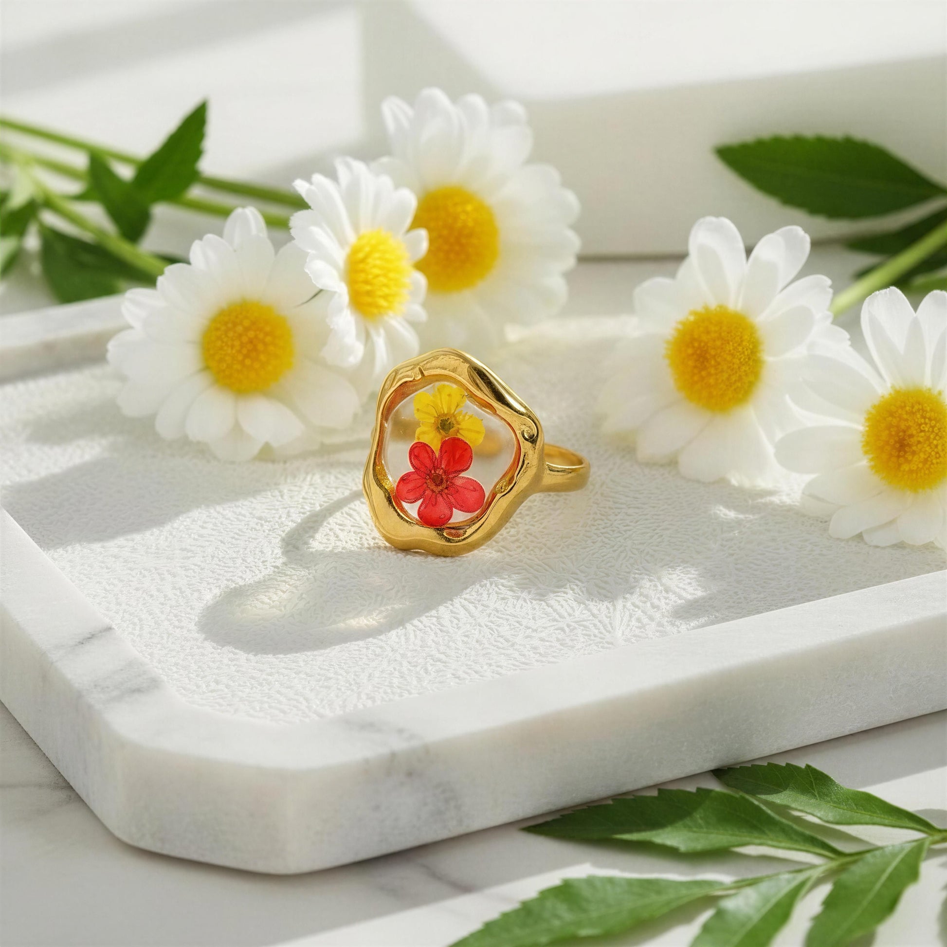 Gold ring with a red flower inset on a marble surface surrounded by white daisies.