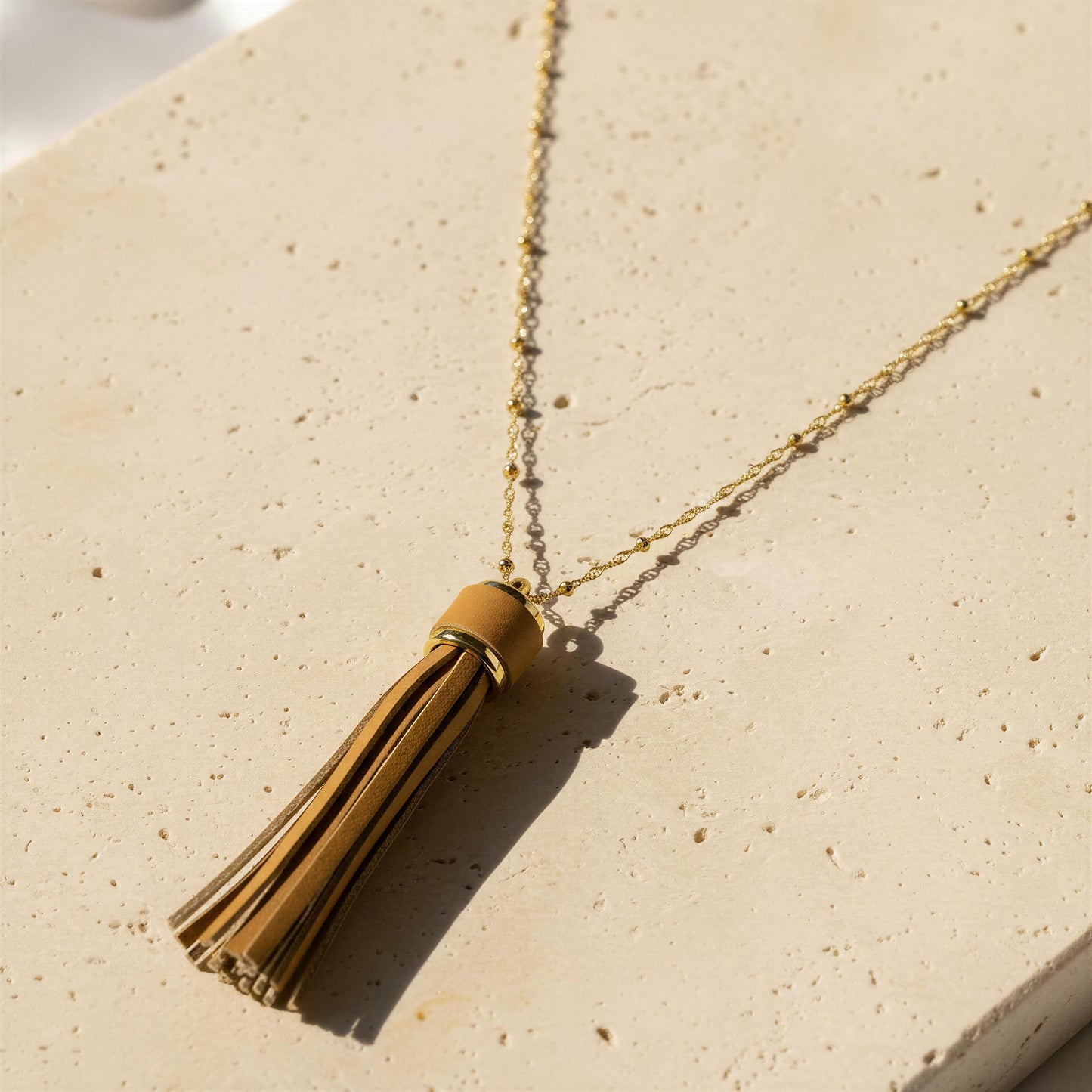 A 18k gold plated beaded chain necklace with a tan leather tassel pendant, styled on a textured beige stone surface.