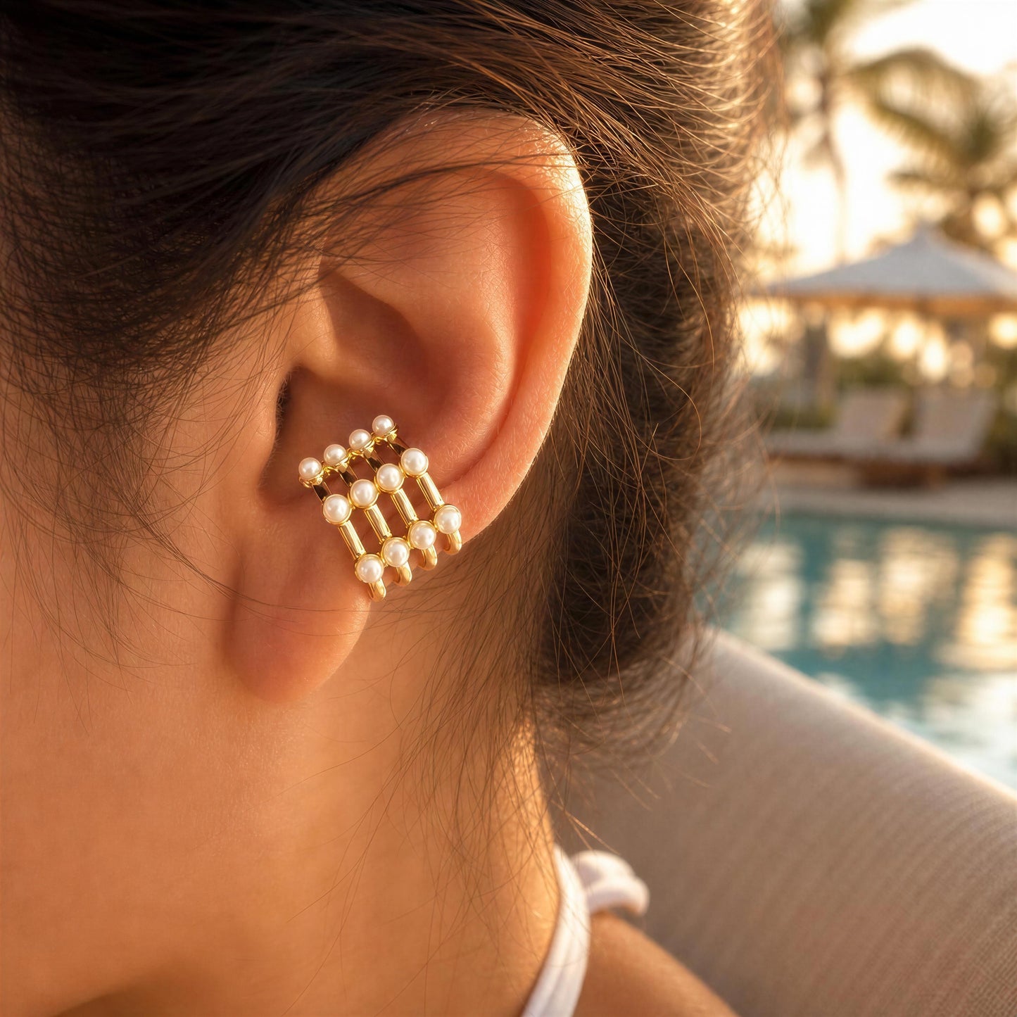 Close-up of an ear with a gold and pearl earring, pool and cabana in the background