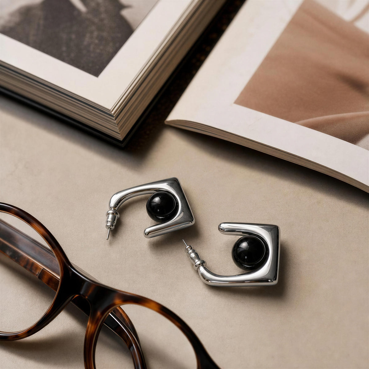 A pair of silver-toned geometric hoop earrings with black oval centers, styled on a beige tabletop with open books and tortoiseshell glasses nearby.