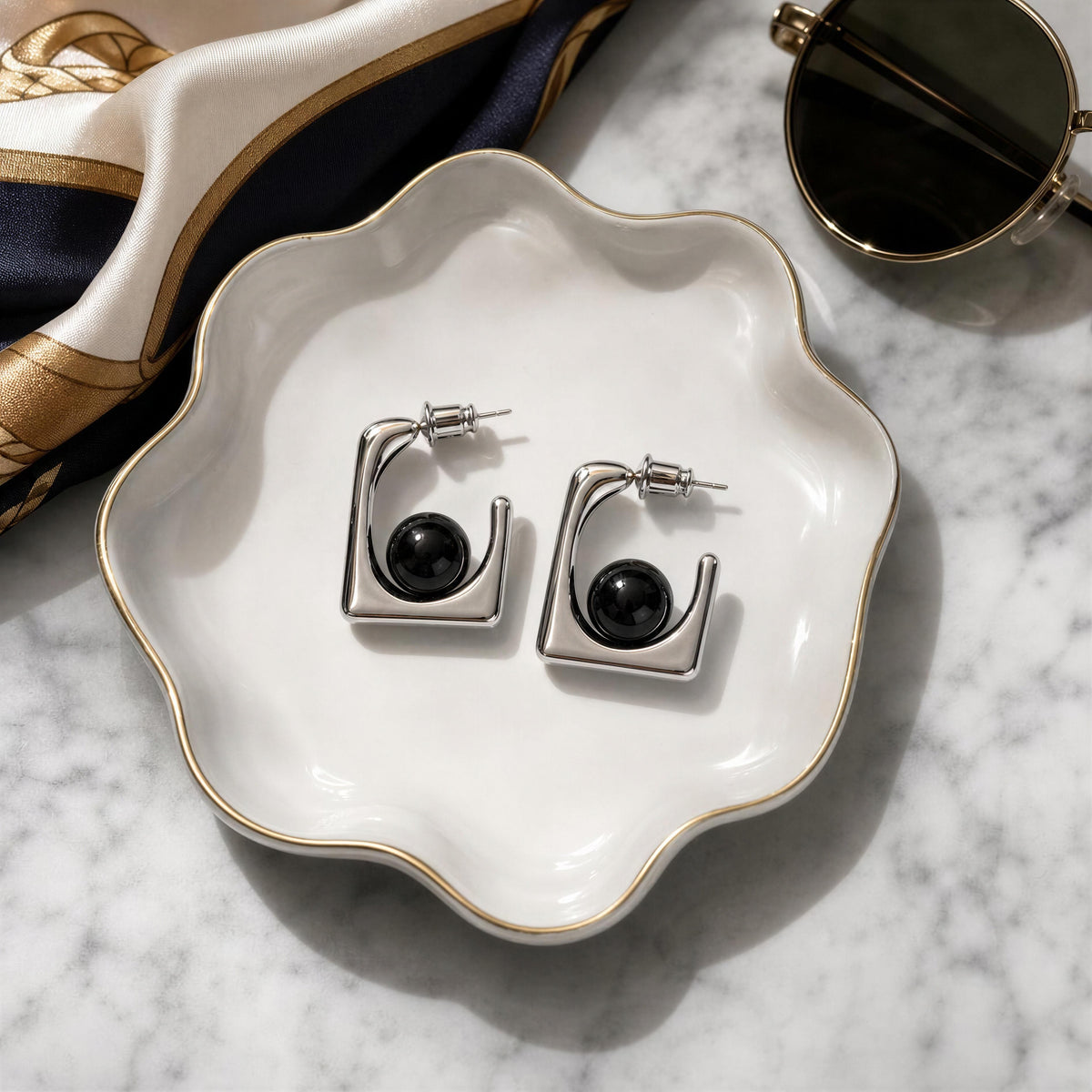 A pair of silver-toned geometric hoop earrings with black oval centers, displayed on a white scalloped dish over a marble surface with a silk scarf and sunglasses nearby.