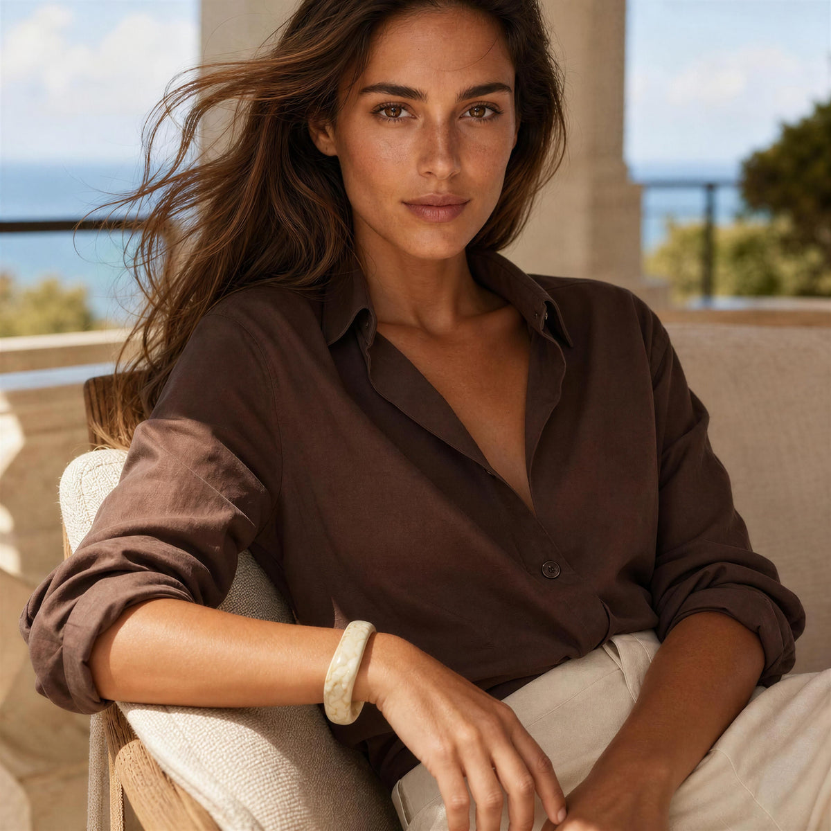 A woman wearing a dark brown linen button-down shirt and off-white trousers, sitting on a beige outdoor lounge chair with a blurred ocean background and the Krem Bangle from Chica Chica Jewelry.