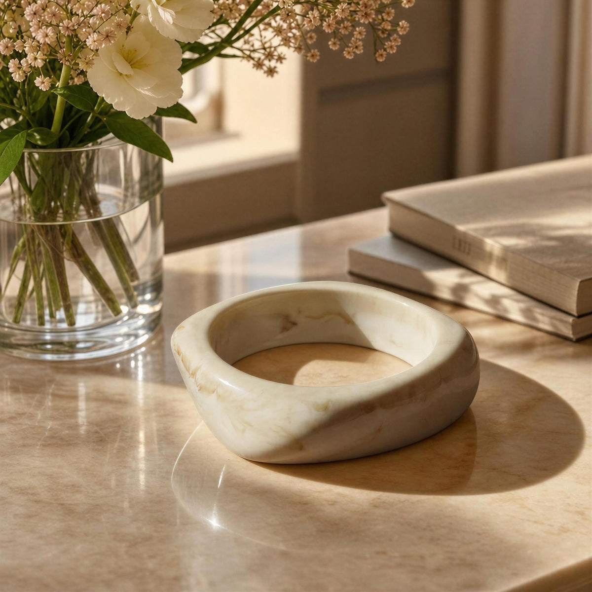 The Chica Chica Jewelry Krem Bangle. A round, chunky bangle bracelet made of cream-colored marble with soft beige and light brown veining, displayed on a polished marble tabletop next to a glass vase of white flowers and stacked books.
