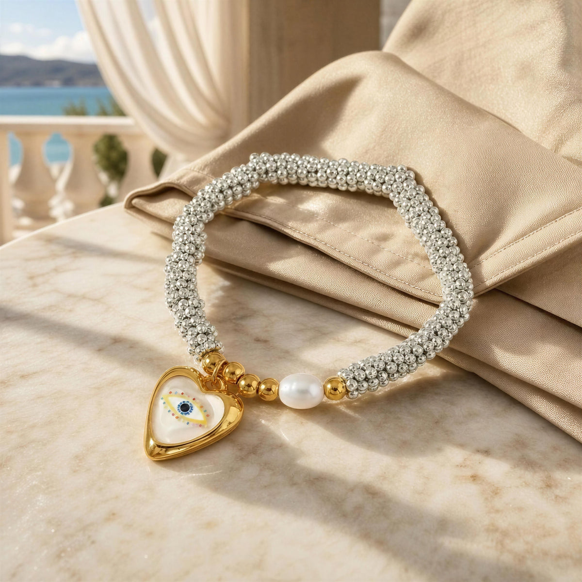 A beaded bracelet with a gold heart charm featuring an evil eye motif, resting on a beige satin fabric draped over a marble surface.