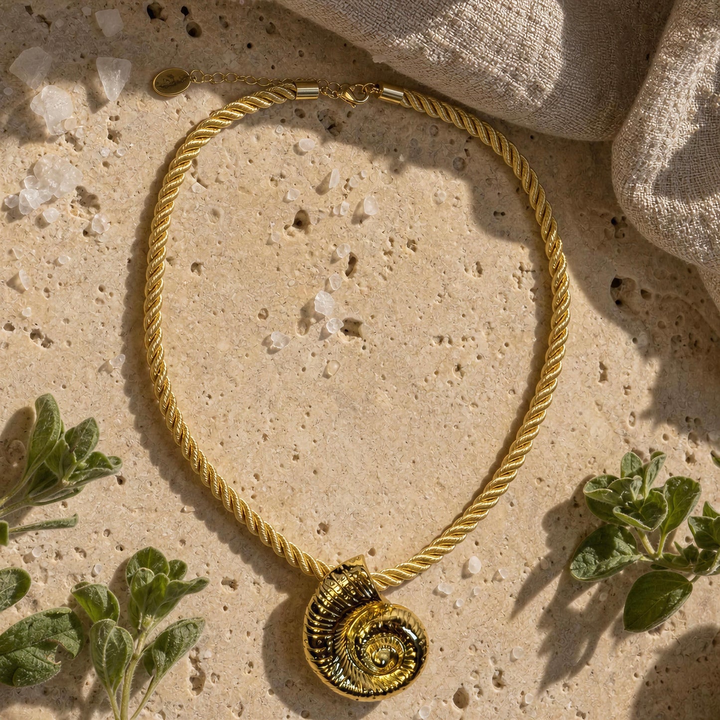 Th  Nautila Necklace from  Chica Chica Jewelry  with a spiral shell pendant, styled on a textured beige stone surface with small green leaves and a linen cloth in the background.