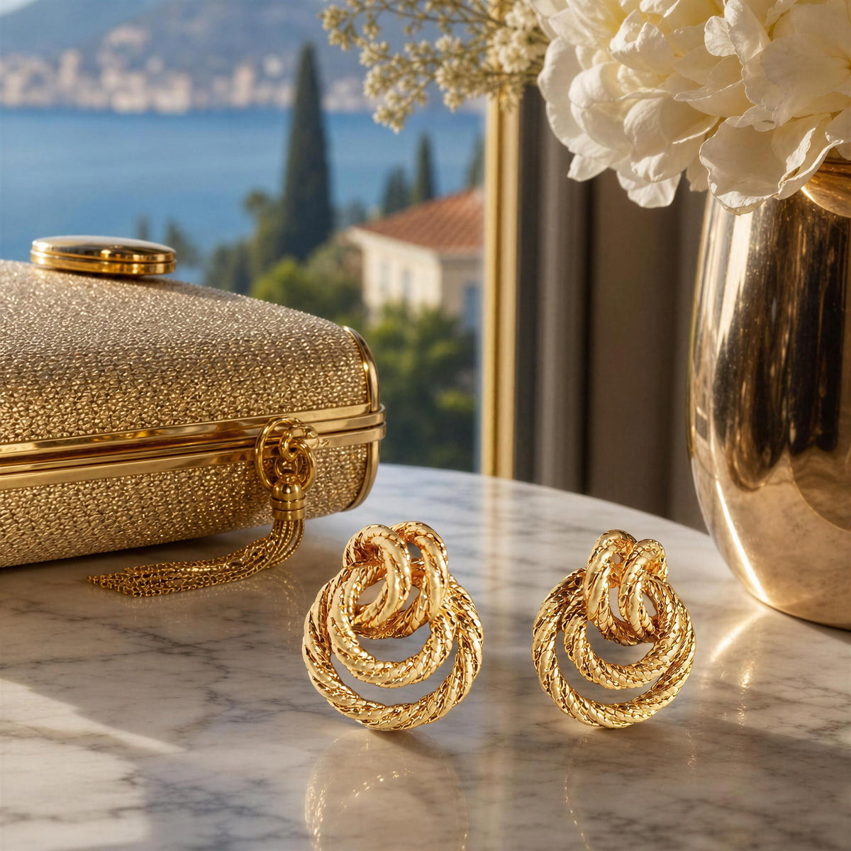 A pair of gold knotted hoop earrings displayed on a marble surface next to a textured gold clutch, with a vase of white flowers and a scenic window view in the background.