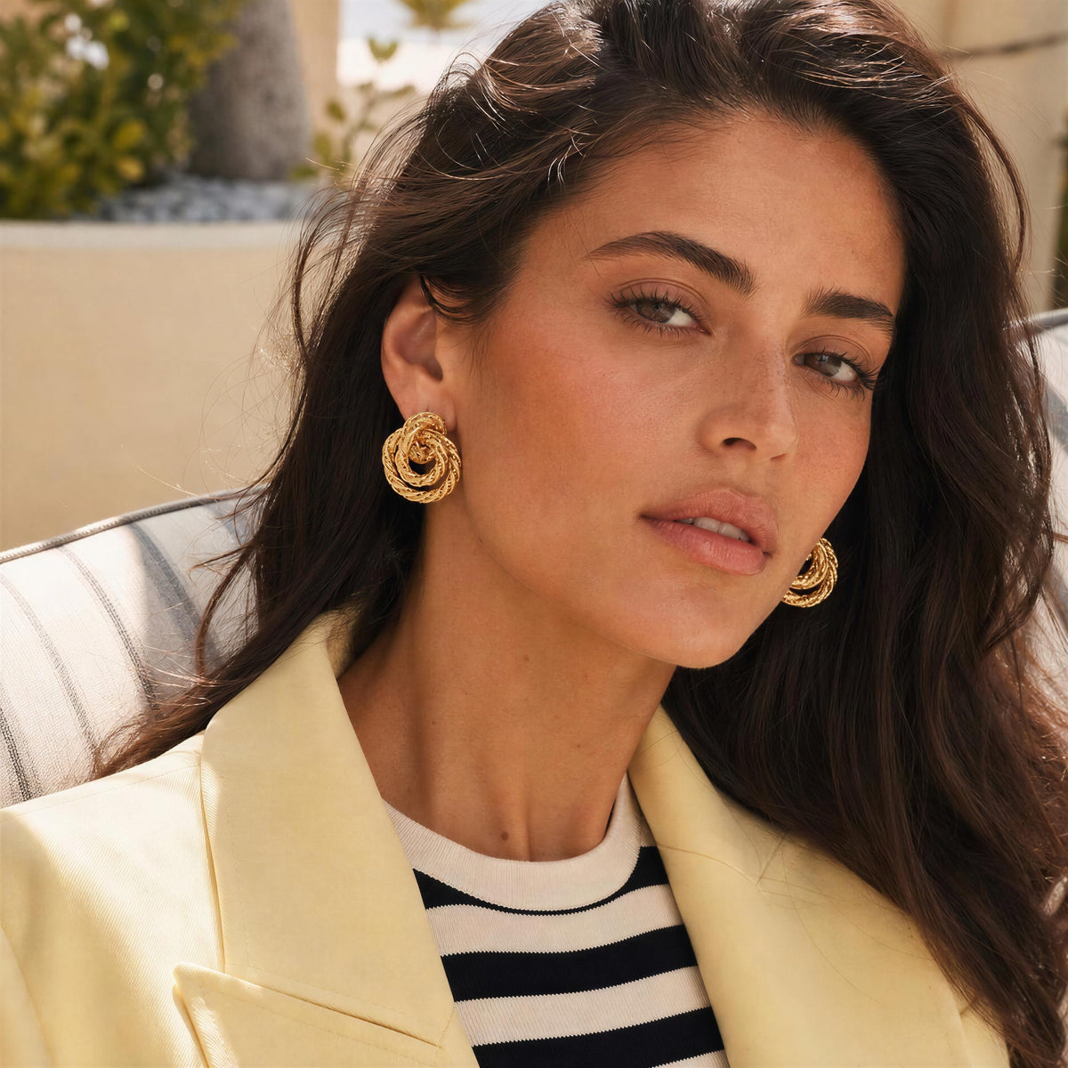 A close-up lifestyle shot of a woman wearing the  Chica Chica Jewelry Nodo Earrings, textured gold spiral drop earrings, a cream blazer, and a black and white striped shirt