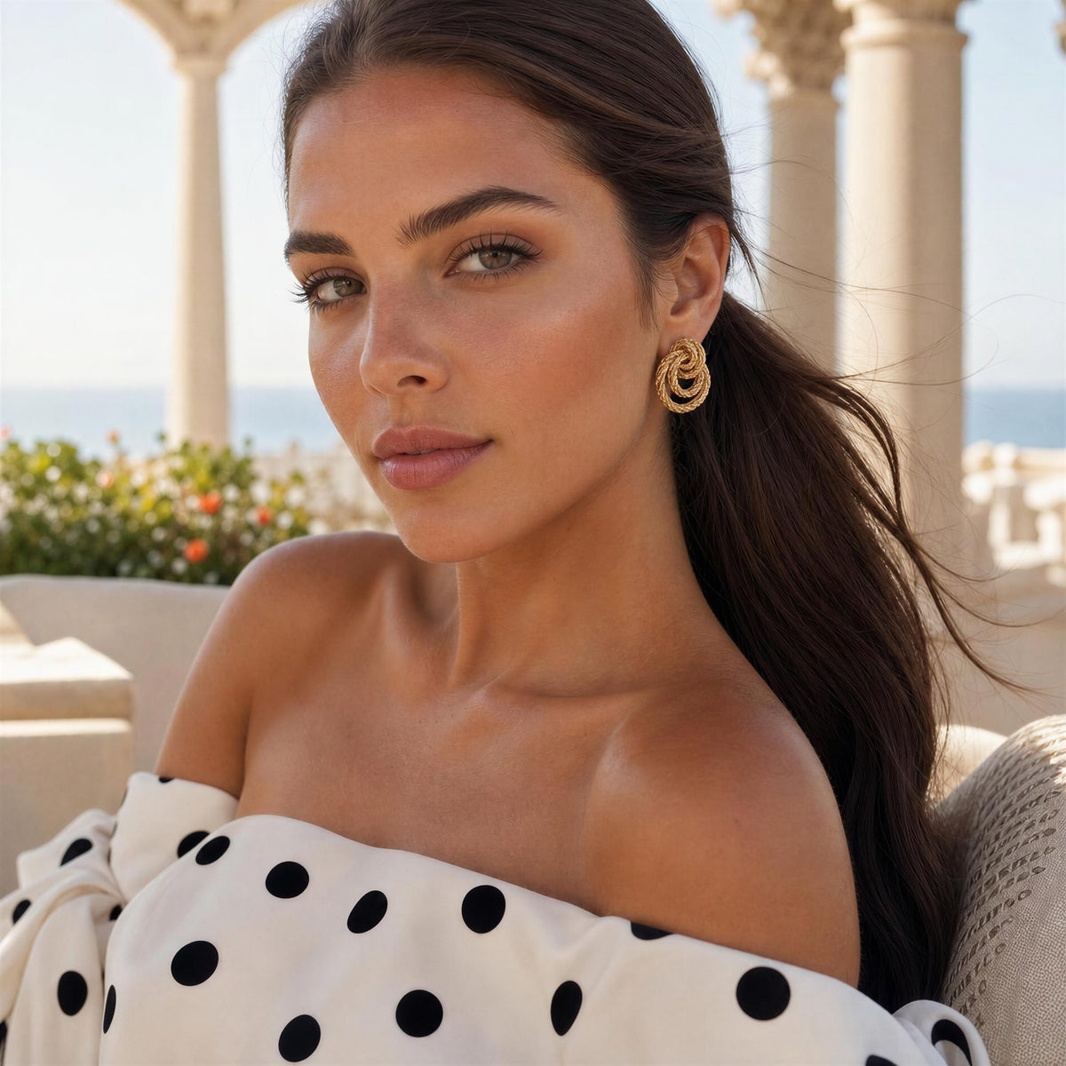 A woman wearing a pair of gold-toned spiral drop earrings, styled with a white off-the-shoulder top featuring black polka dots, set against a sunny outdoor patio background with columns and the sea in the distance.