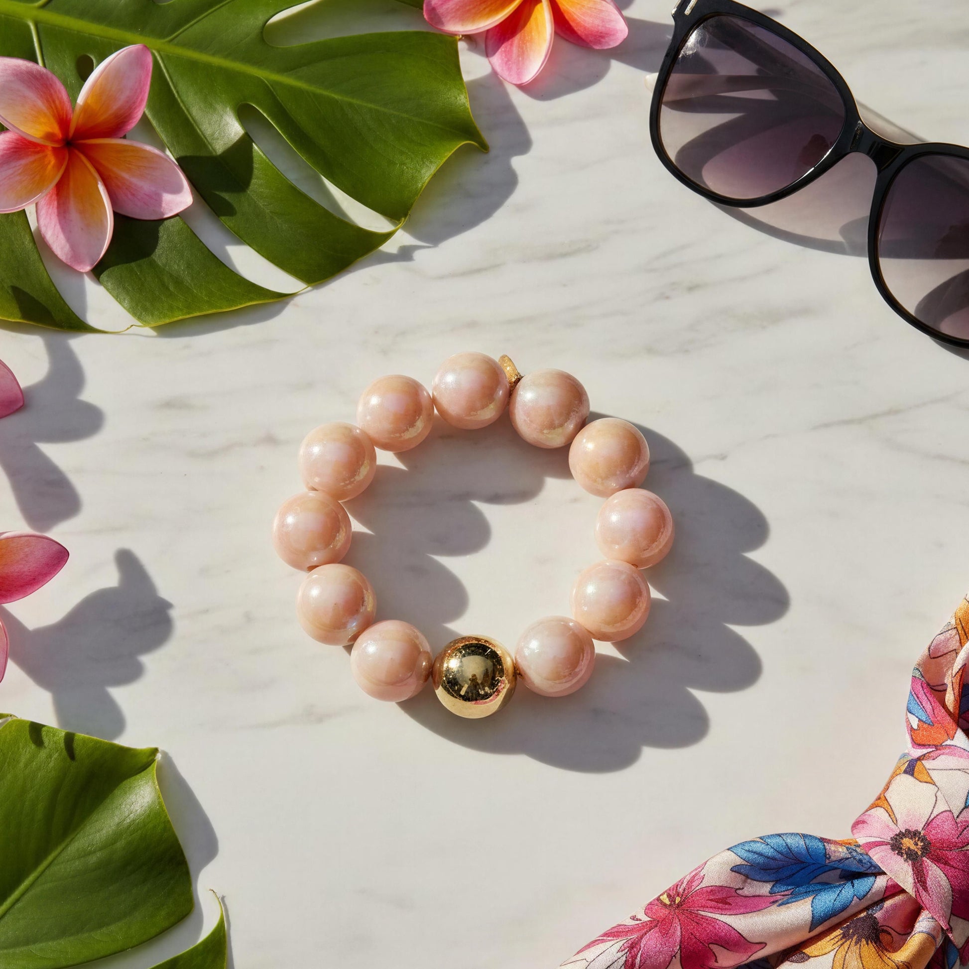 Peach beaded bracelet with a gold charm on a white surface with flowers and sunglasses.